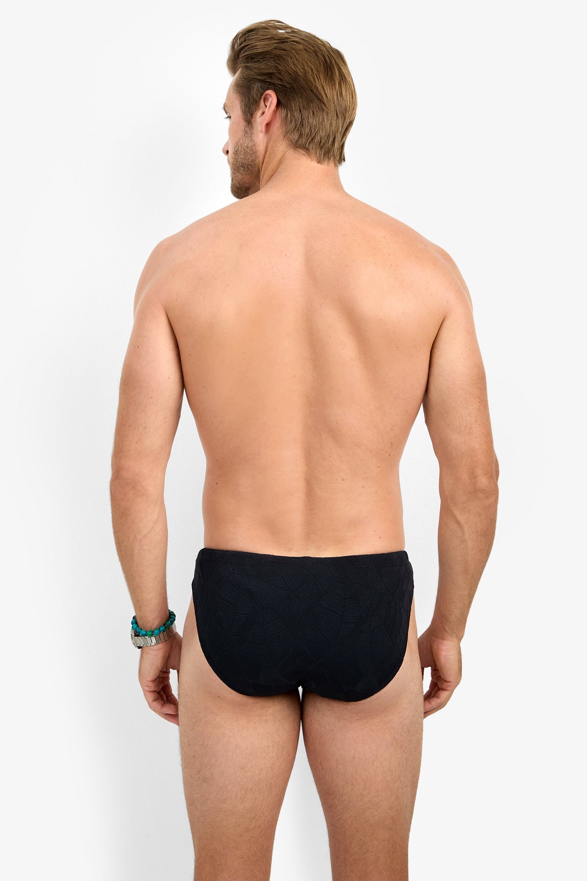 A man with light skin and short, light brown hair stands facing away against a plain white background, wearing only the LAGOS SWIM BRIEF in black and a turquoise beaded bracelet on his left wrist.