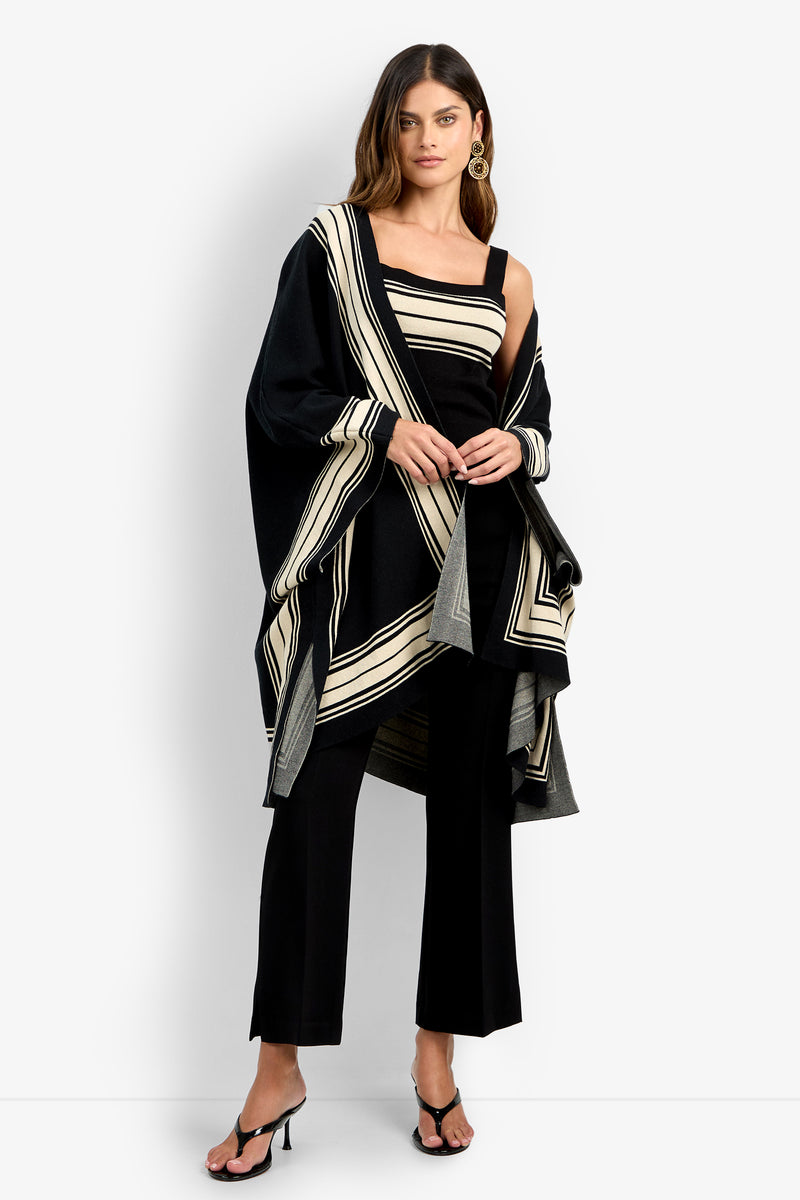A woman wears the VILLAS PONCHO, a stylish black and beige striped shawl, paired with wide-leg pants, a matching top, hoop earrings, long hair, and black open-toe heels.