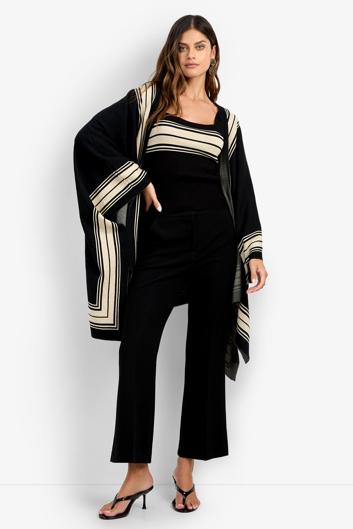 A woman poses with one hand on her hip against a plain light background, wearing the VILLAS PONCHO—a black outfit featuring cream geometric stripes, styled with a matching tank top, black pants, and heeled sandals.