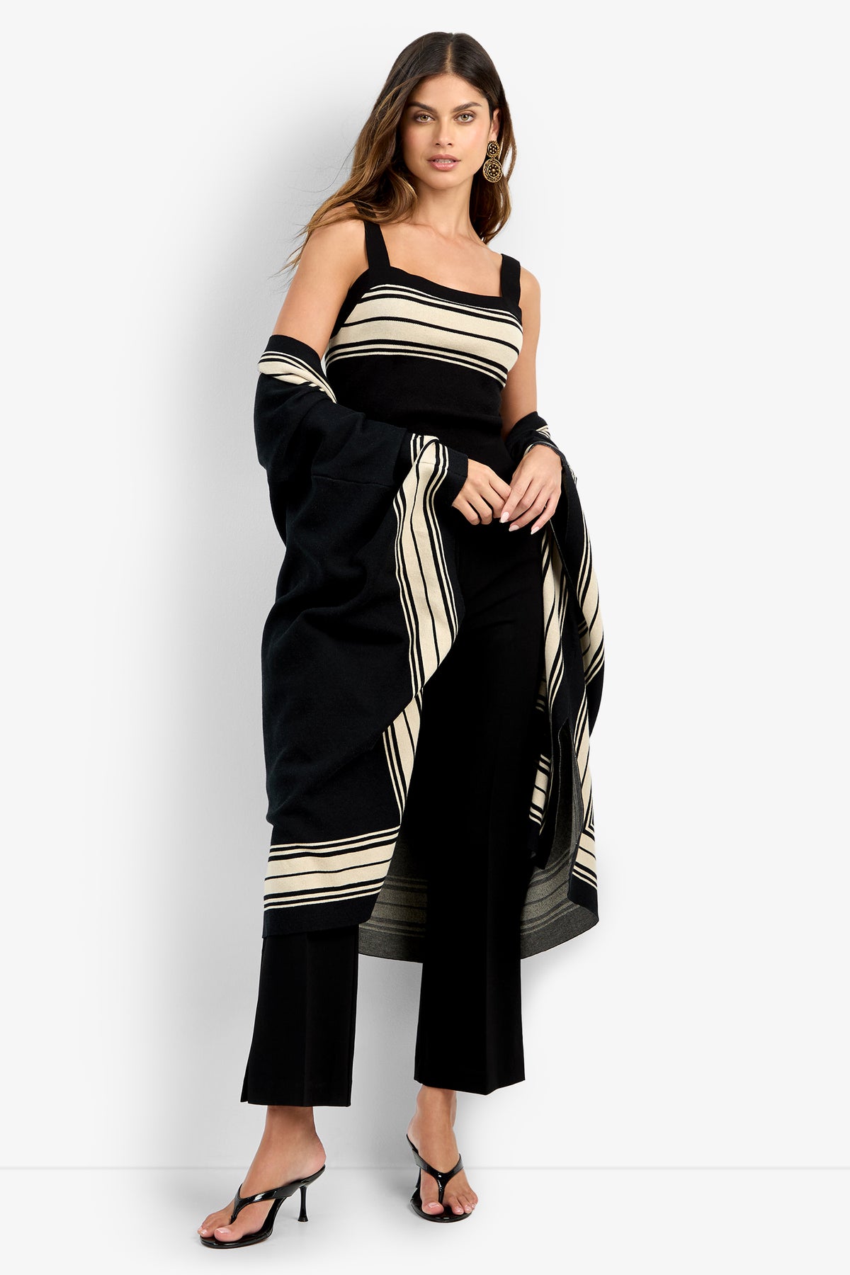 A woman with long brown hair and gold hoop earrings stands against a white background wearing the VILLAS PONCHO, styled with black pants and black heeled sandals.