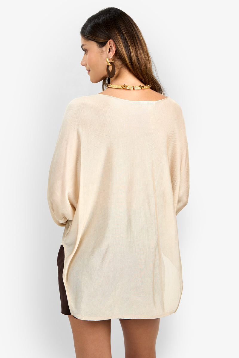 A woman with long brown hair, gold hoop earrings, and a chunky gold necklace faces away, wearing the VINO PULLOVER—a loose, long-sleeve beige blouse—over brown shorts against a plain light background.