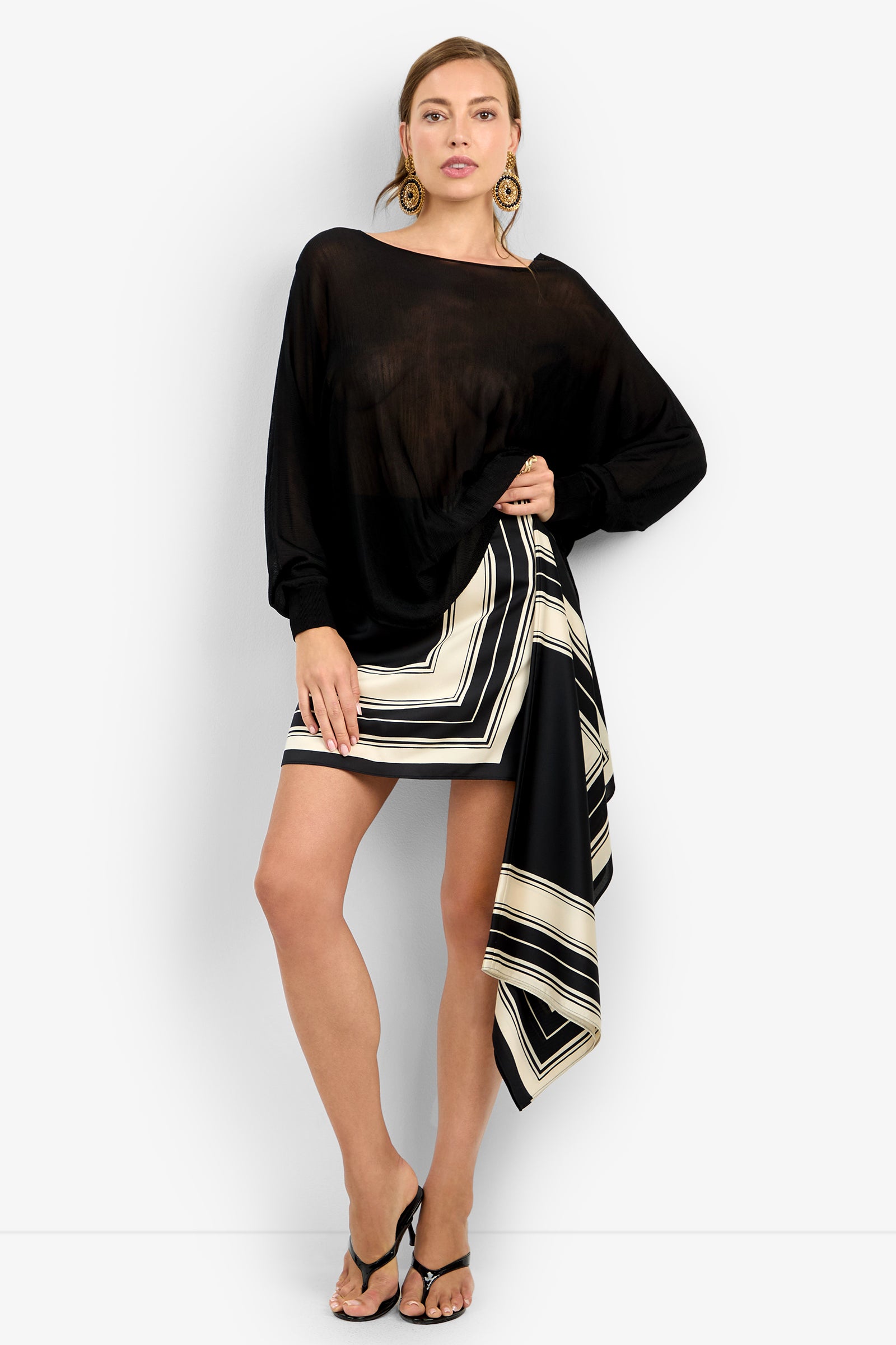 Woman stands against a white background wearing the VINO PULLOVER, a sheer black long-sleeve top, paired with a geometric black, white, and beige asymmetrical skirt, black heeled sandals, and large gold earrings.