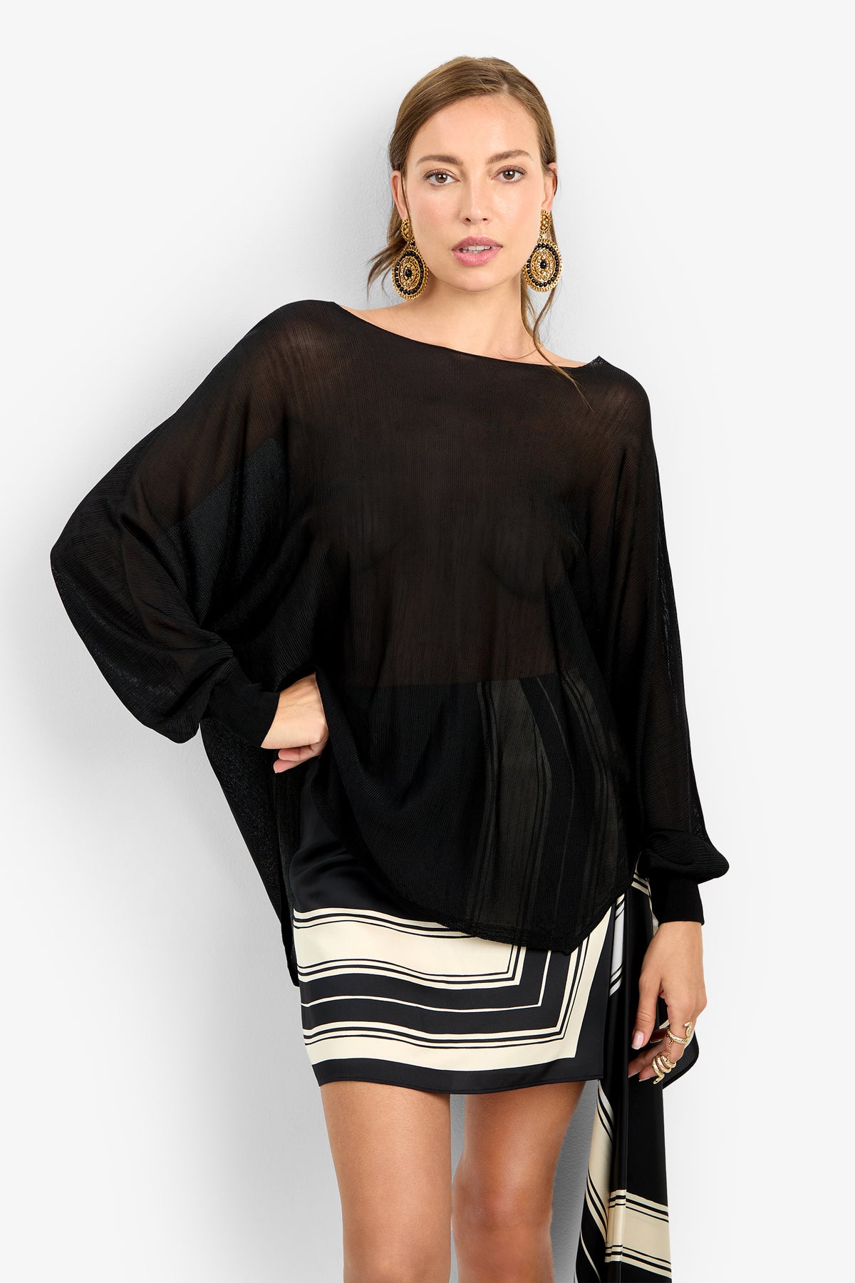 Model wears the VINO PULLOVER, a loose, sheer black long-sleeve top, over a black and white geometric-patterned skirt. She poses against a plain white background with light brown hair, large circular earrings, and a neutral expression.