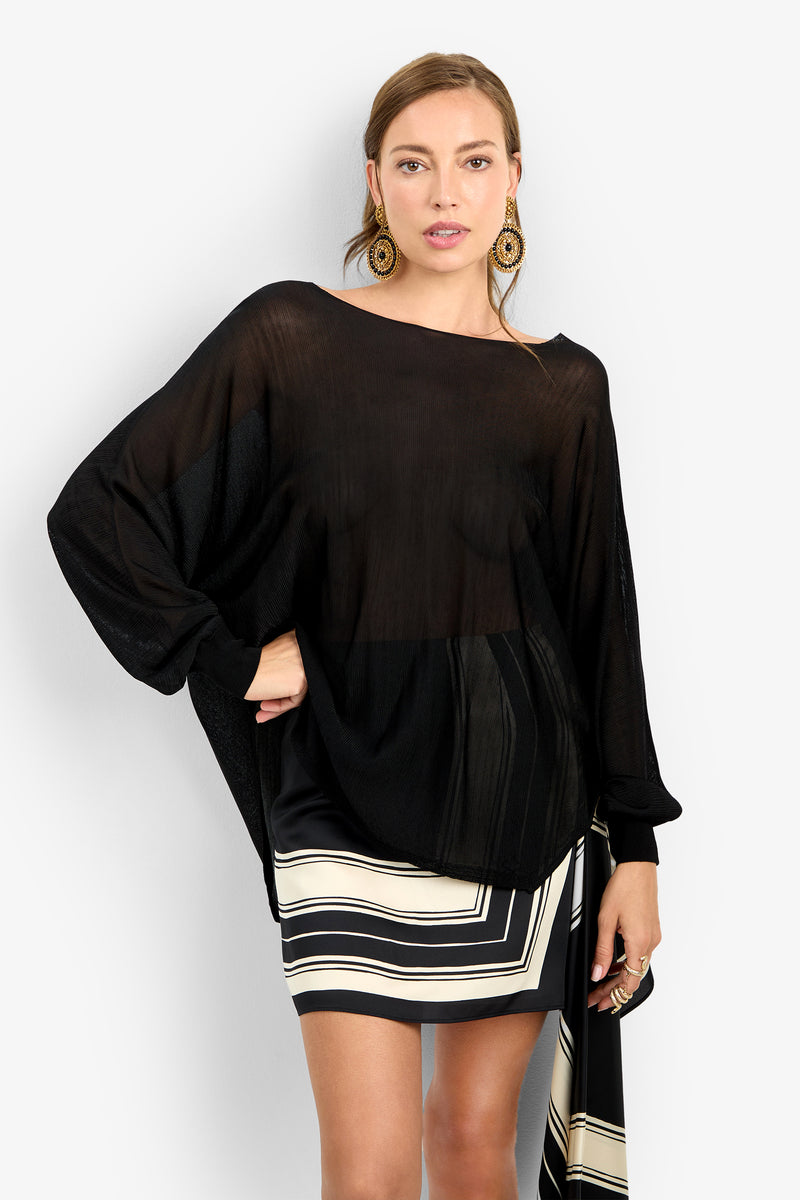 Model wears the VINO PULLOVER, a loose, sheer black long-sleeve top, over a black and white geometric-patterned skirt. She poses against a plain white background with light brown hair, large circular earrings, and a neutral expression.