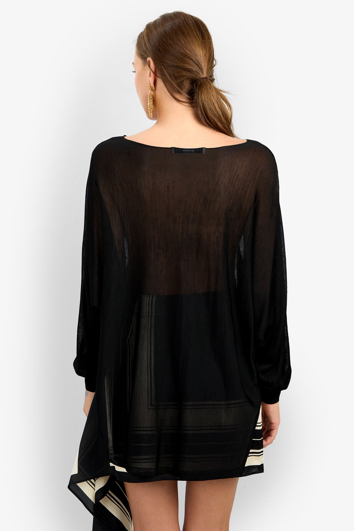 A woman with light brown hair in a low ponytail is seen from the back wearing the VINO PULLOVER, a loose, sheer black top with long sleeves and a square neckline, paired with gold hoop earrings.
