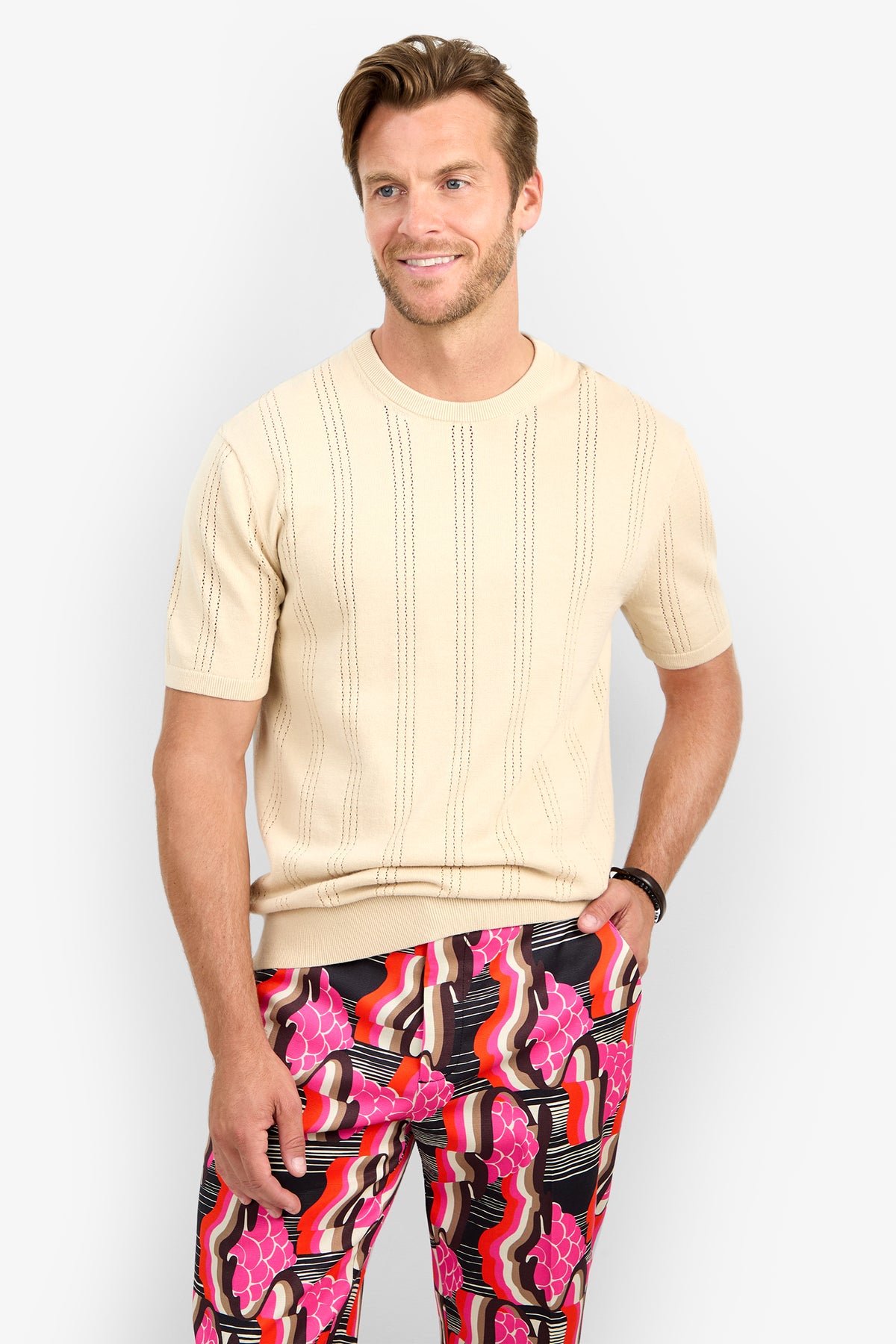 A man with light brown hair and a beard wears the beige CARLO CREWNECK short-sleeve sweater and colorful, patterned pants with red, pink, black, and white designs. He stands smiling against a plain white background.