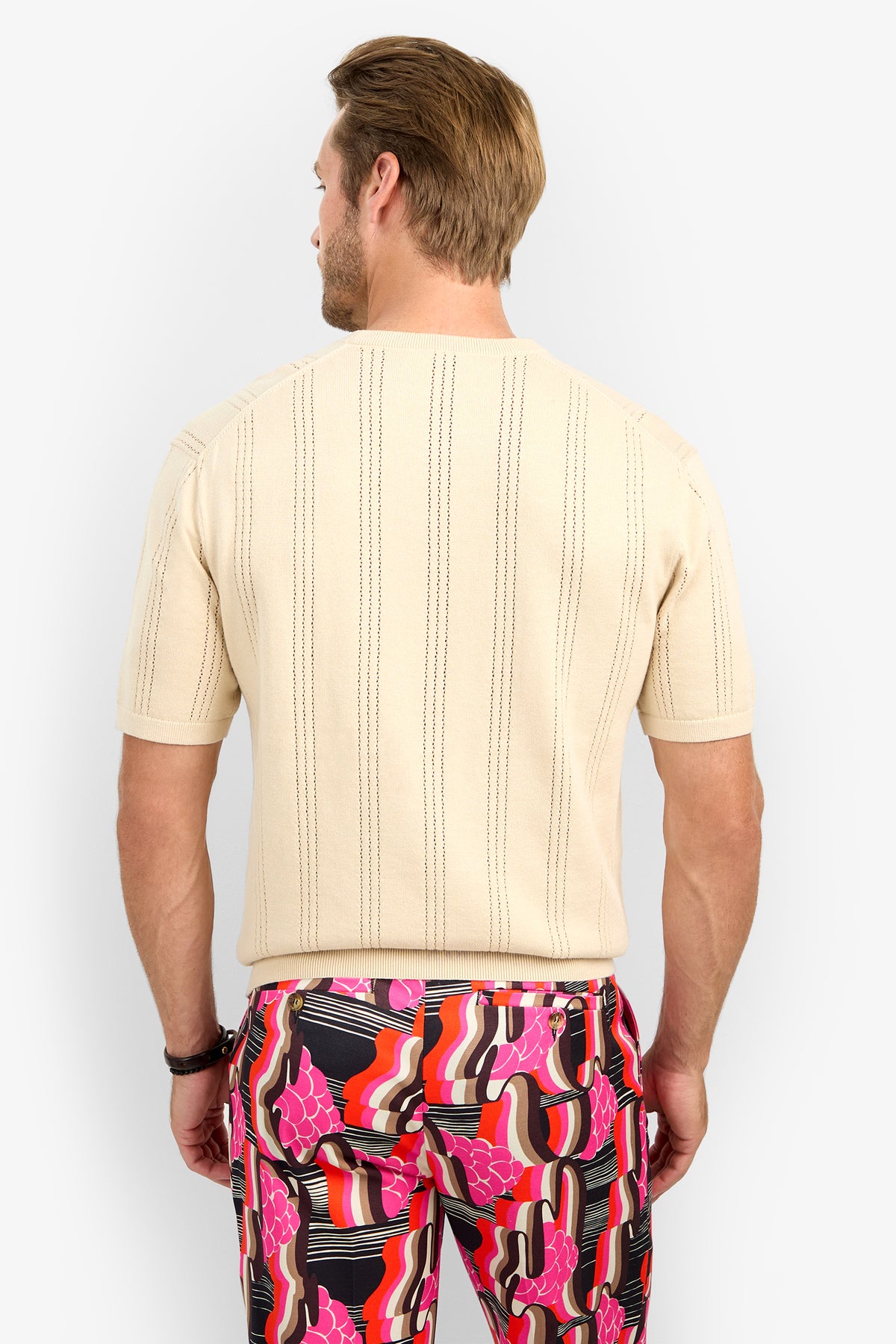 A man is seen from behind wearing the CARLO CREWNECK—a short-sleeved, beige textured shirt—paired with bold pants featuring a pink, red, black, and white abstract pattern.