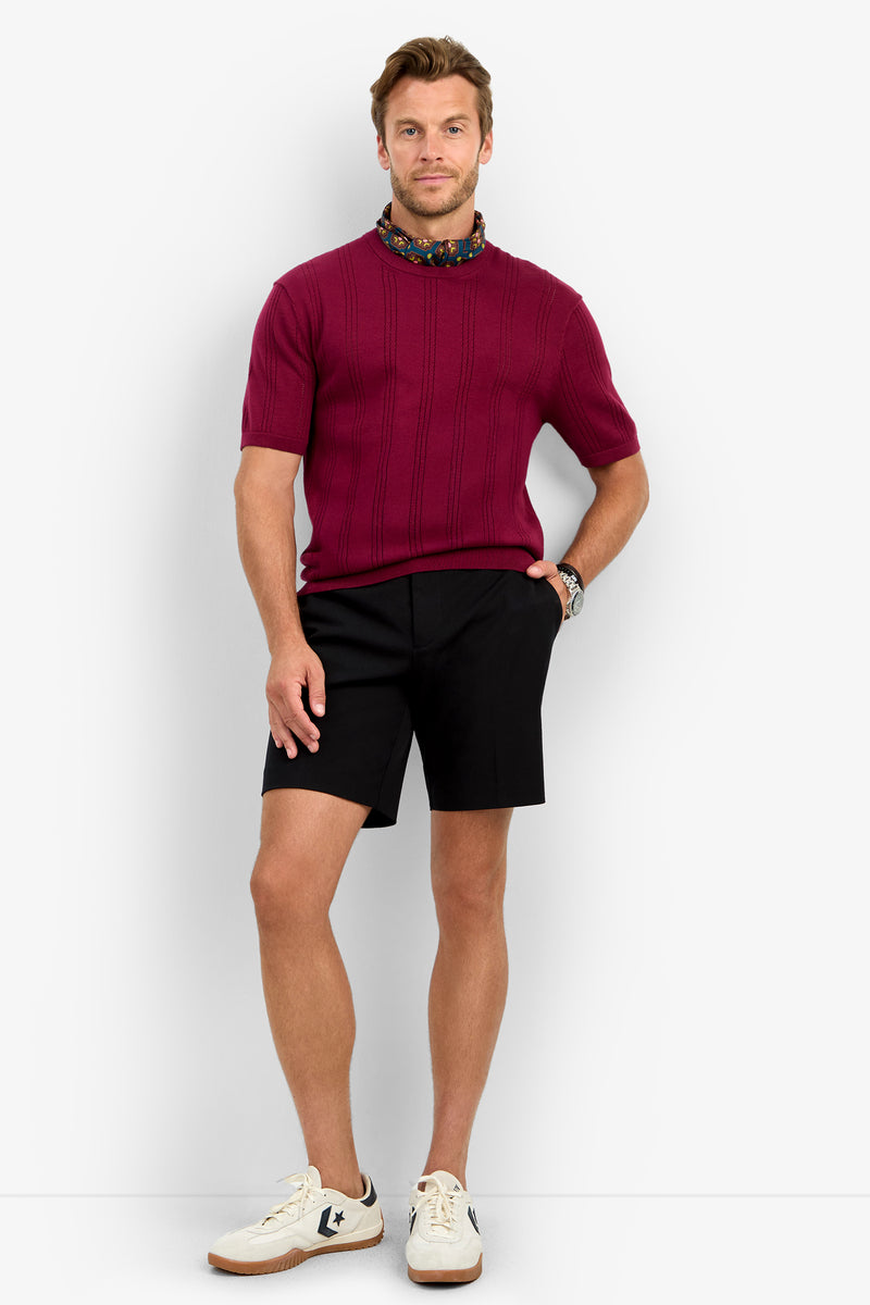 Wearing the CARLO CREWNECK short-sleeve burgundy sweater, a man with light brown hair pairs it with black shorts, white sneakers, a colorful scarf, and a watch on his left wrist against a plain background.