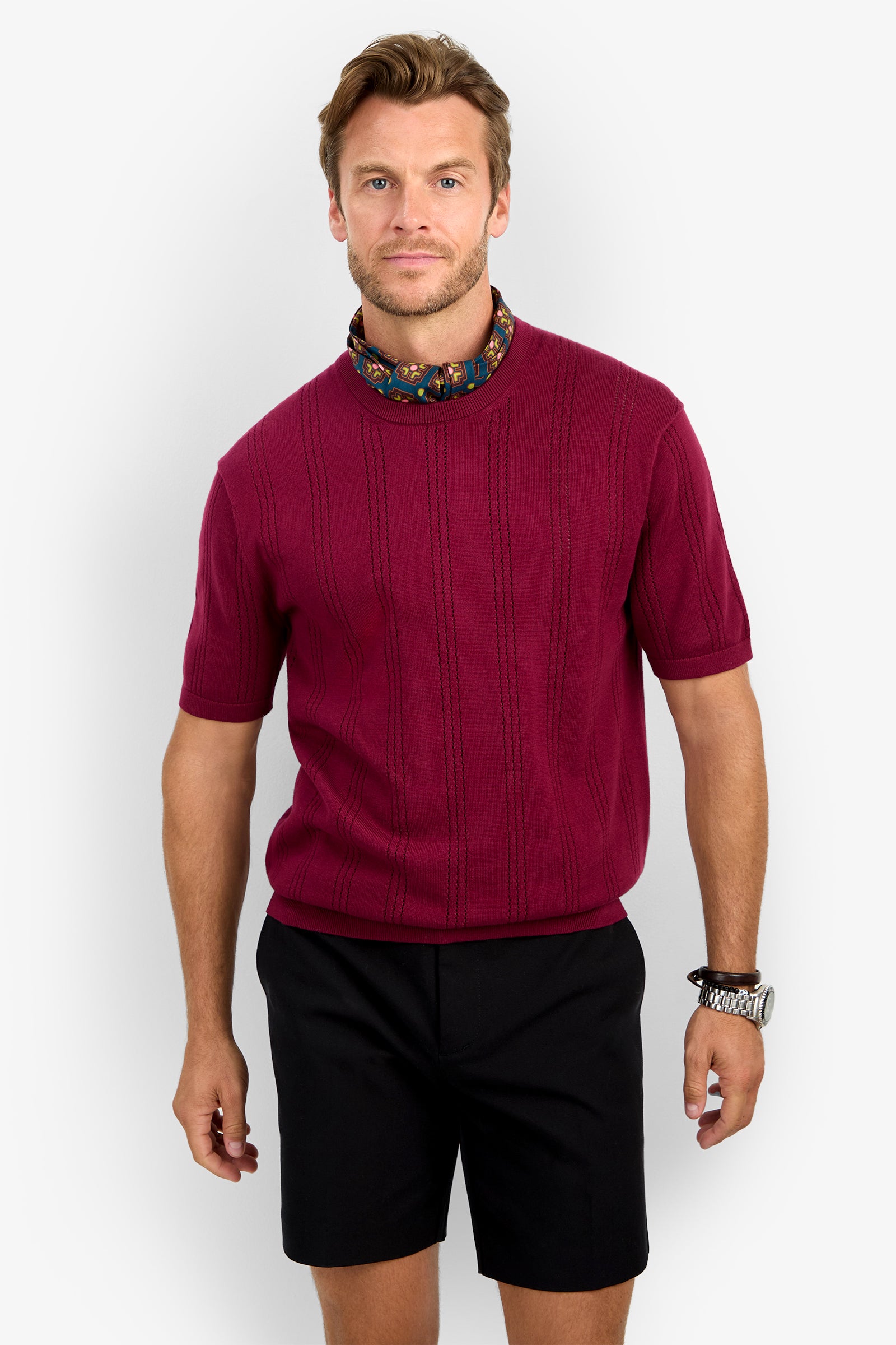A man with light brown hair and facial hair stands against a plain background, wearing the CARLO CREWNECK short-sleeve burgundy sweater, a patterned neck scarf, black shorts, and a silver wristwatch. He has a neutral expression.