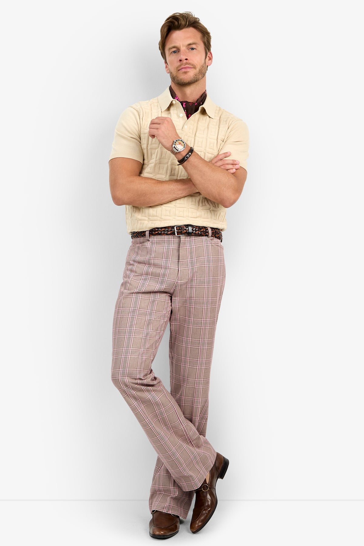 A man stands confidently against a plain white background, arms crossed, wearing the beige VERNON POLO, patterned pink plaid pants, a brown belt, brown shoes, and a wristwatch.