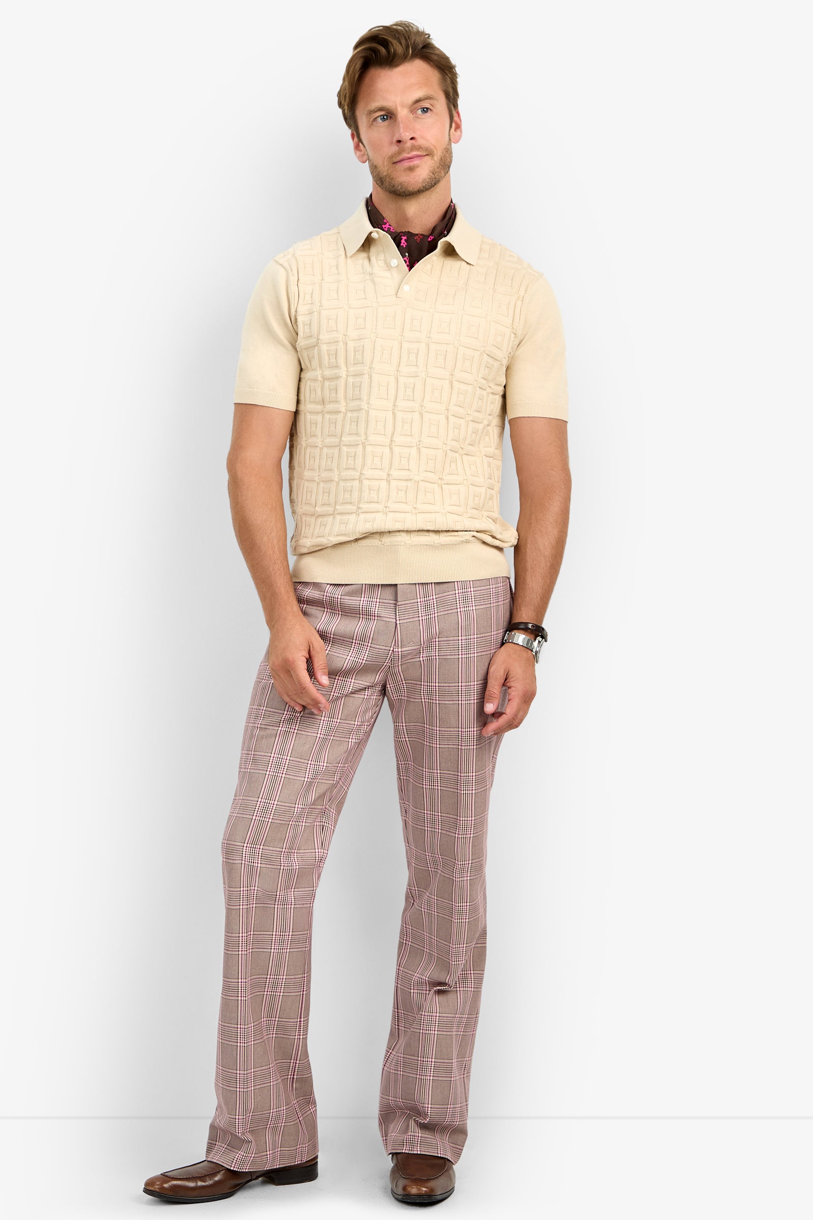 A man stands against a plain background wearing the VERNON POLO, a beige short-sleeve knit sweater, over a maroon collared shirt, pink plaid flare pants, brown dress shoes, and a wristwatch.