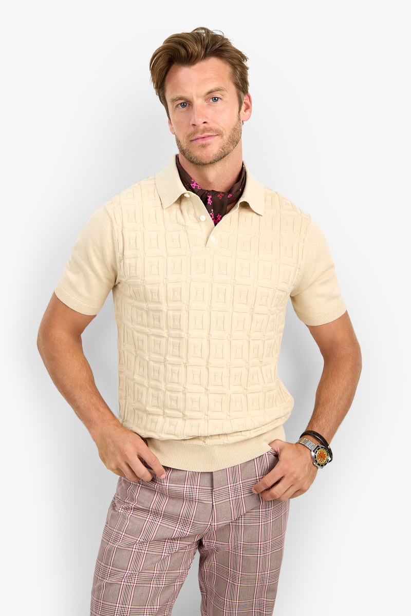 A man wearing the VERNON POLO, a short-sleeve cream textured shirt, along with plaid pants, a patterned neck scarf, and a wristwatch stands against a plain light background with his hands on his hips.
