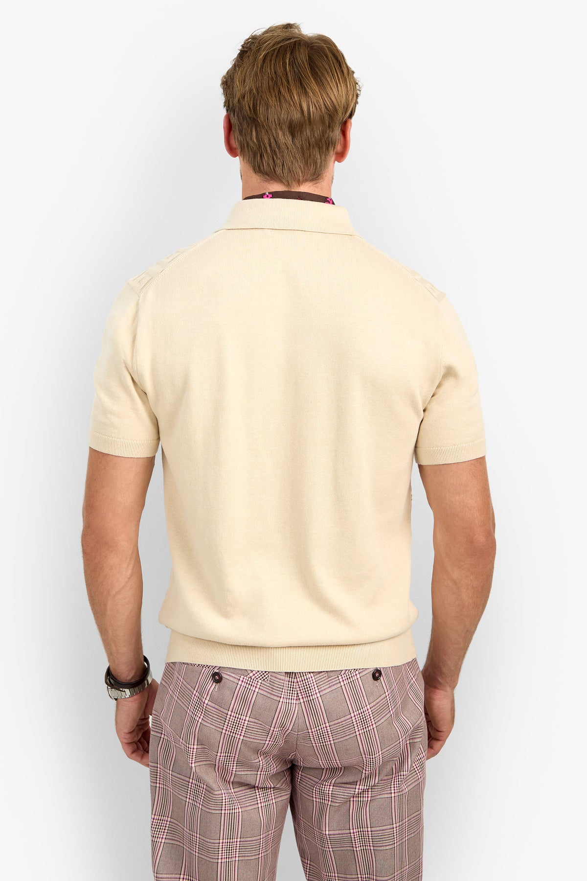 A man with light brown hair is seen from behind, wearing the VERNON POLO in beige and pink plaid pants. He has a watch on his left wrist and stands in front of a plain, light background.
