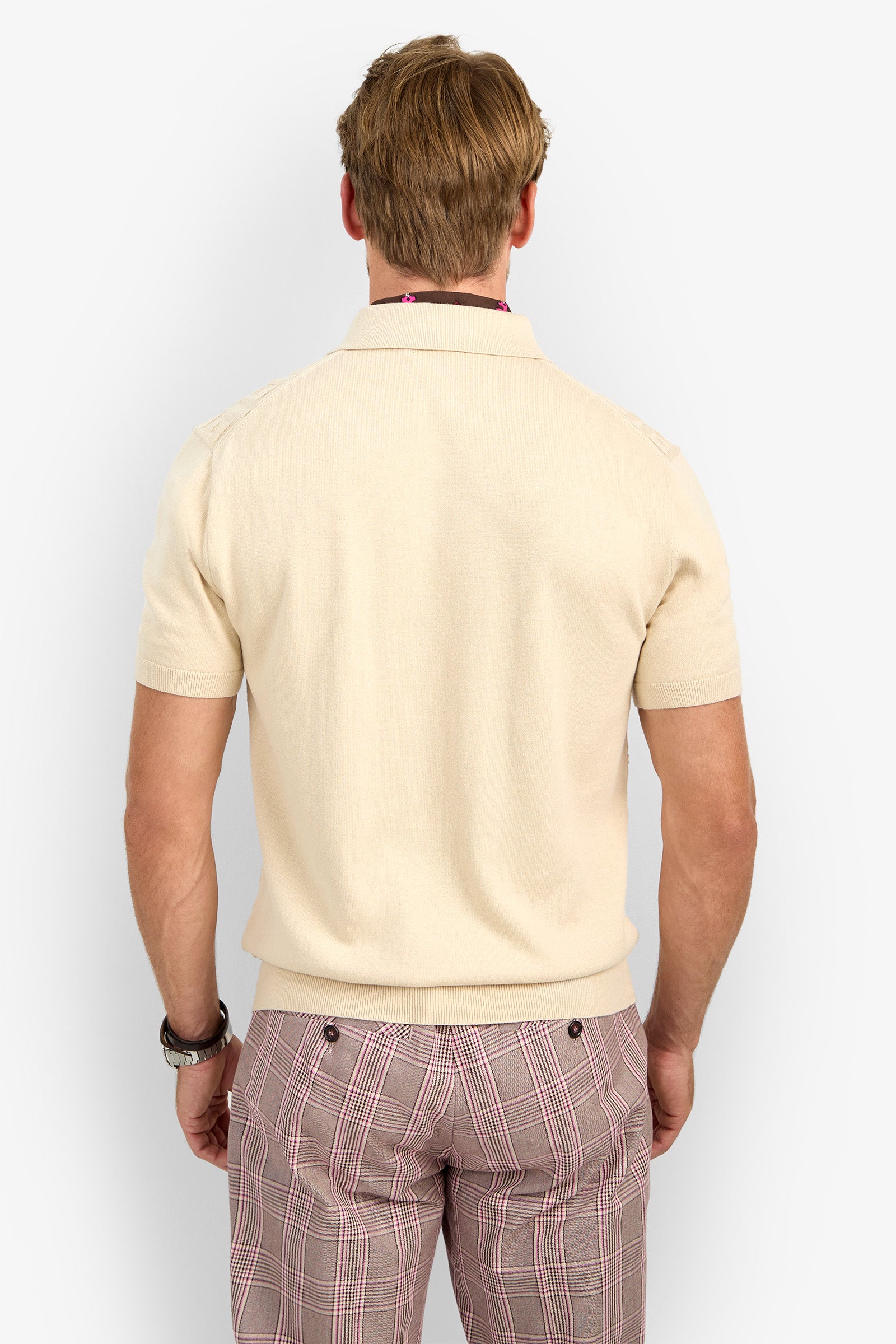 A man with light brown hair is seen from behind, wearing the VERNON POLO in beige and pink plaid pants. He has a watch on his left wrist and stands in front of a plain, light background.