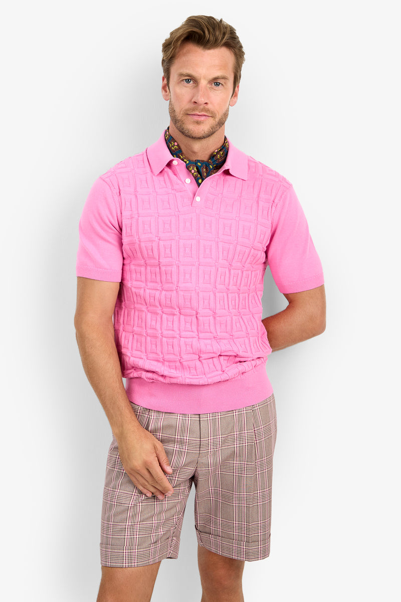 A man wearing the VERNON POLO in textured pink, paired with beige and pink plaid shorts and a vibrant patterned scarf, stands against a plain light background.