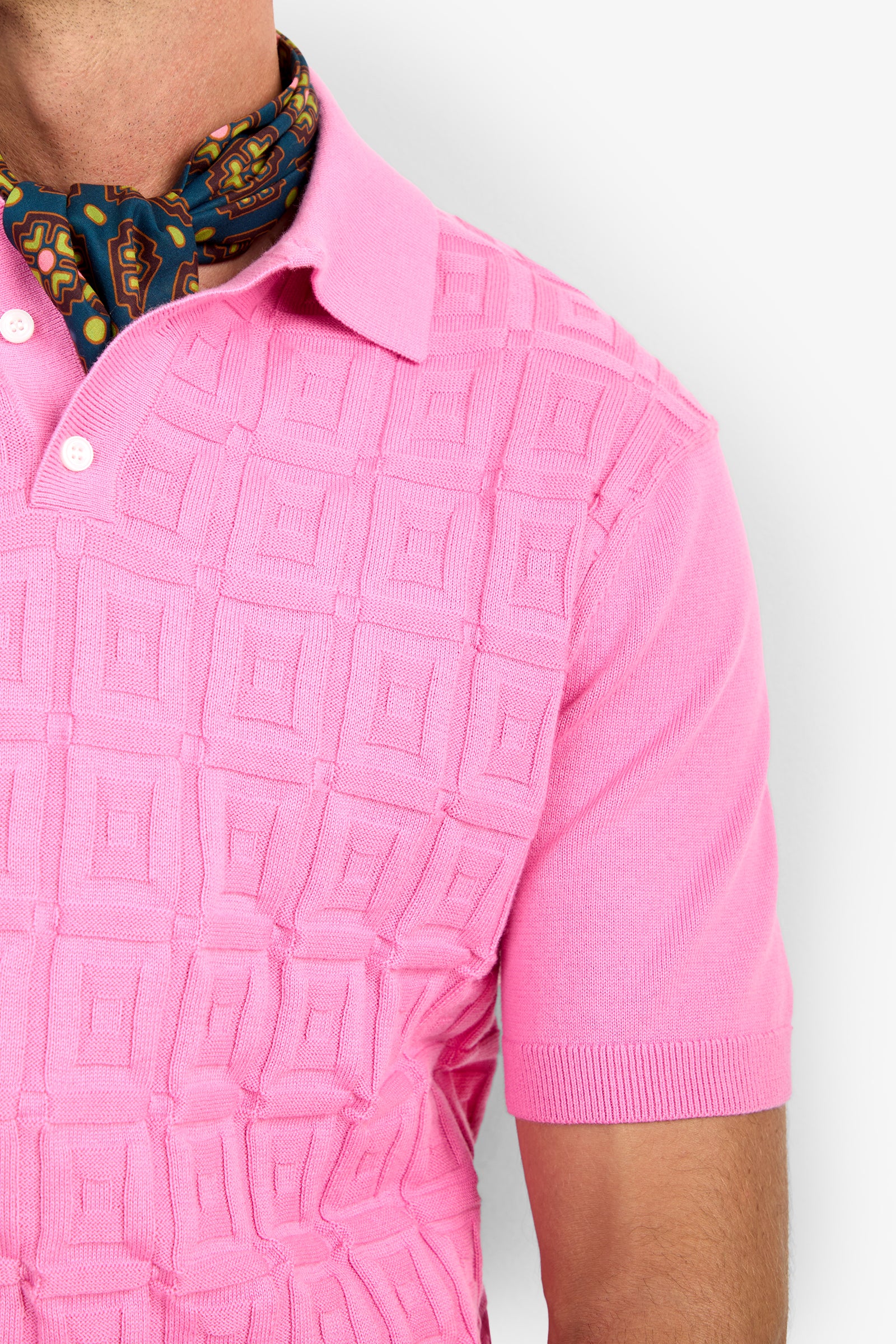 A close-up of a person wearing the VERNON POLO, a textured pink short-sleeve polo shirt with a collar and white buttons, styled with a patterned neck scarf in blue, red, and gold tones.