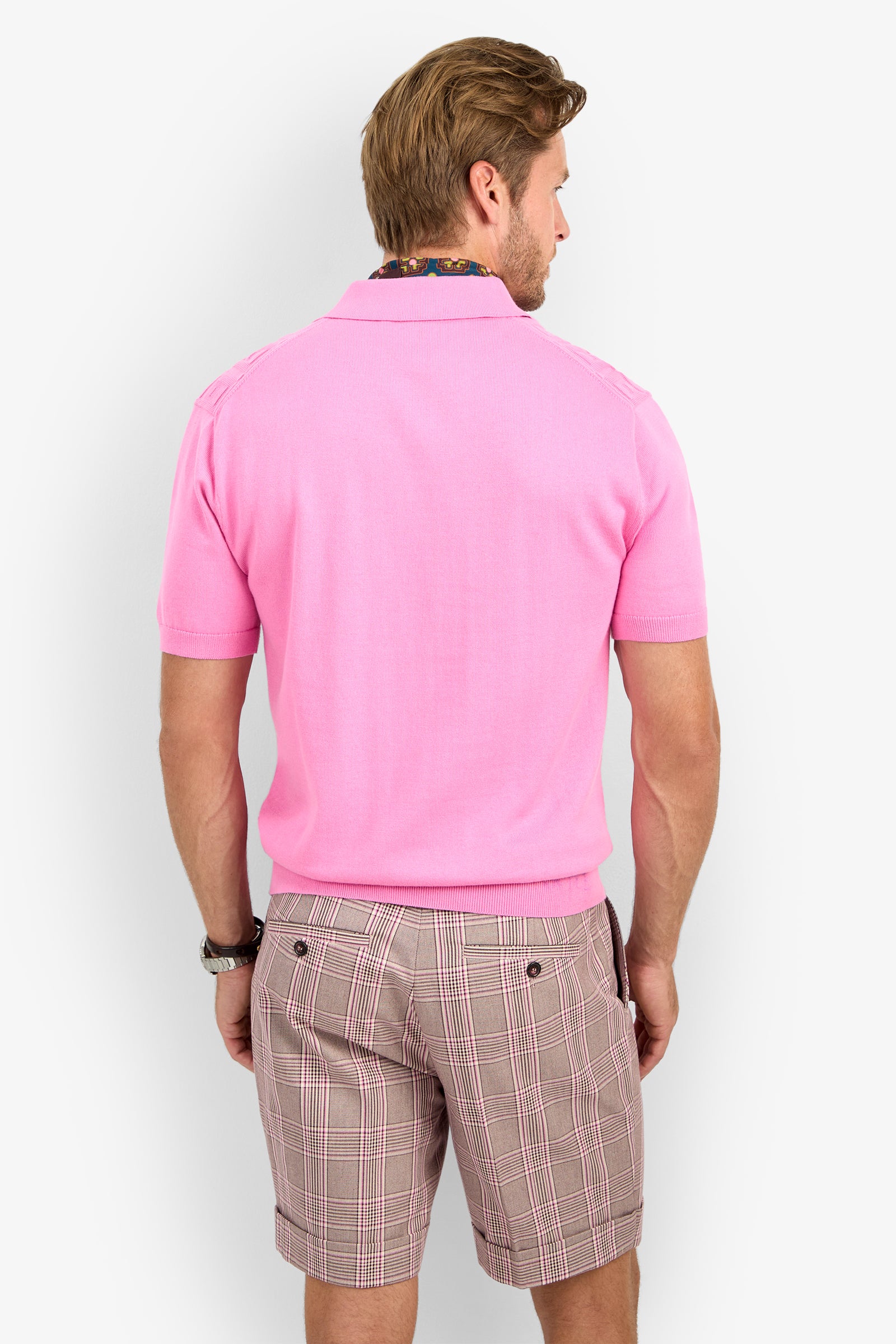 A man with light brown hair faces away, wearing the VERNON POLO in light pink and plaid brown-pink shorts. He has a wristwatch on, set against a plain white background.