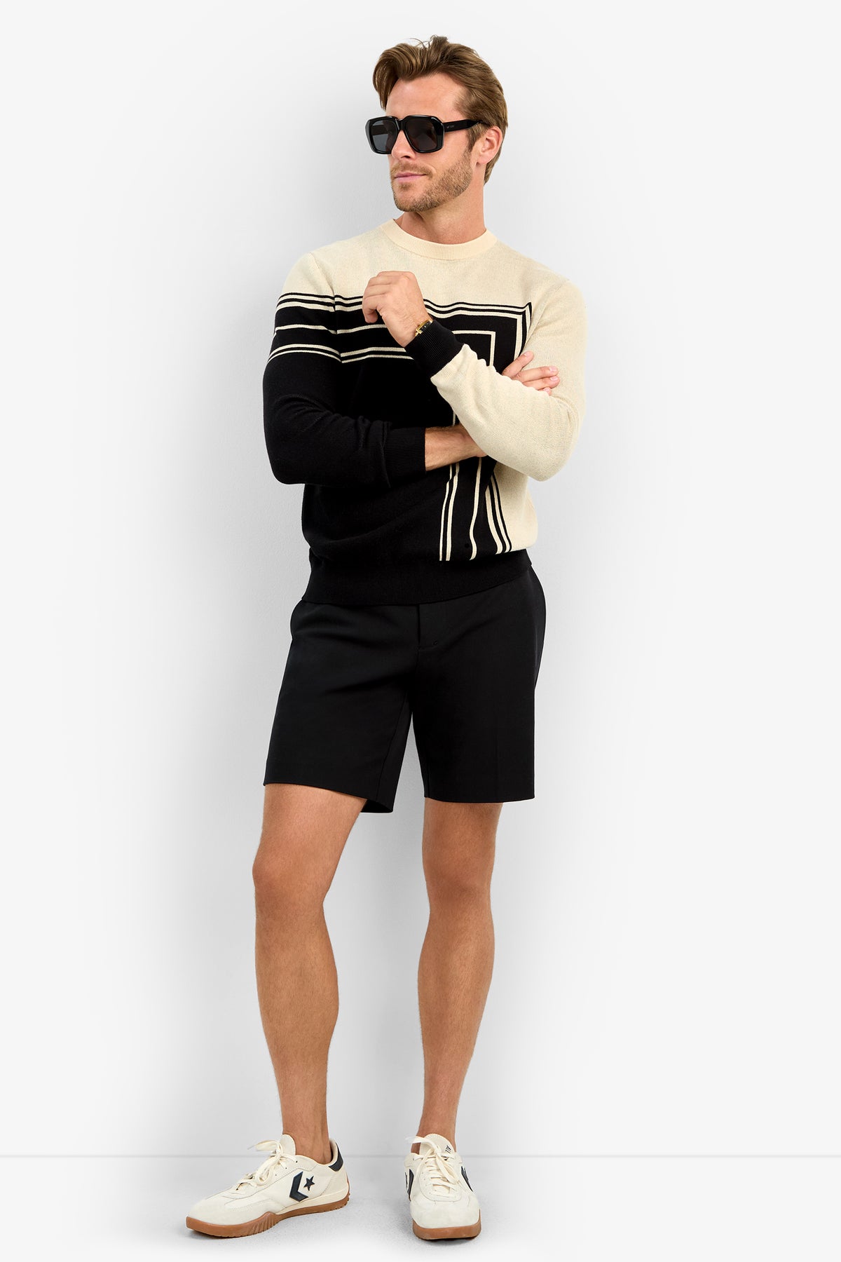 Wearing the RAMOS STRIPE CREWNECK sweater, black sunglasses, black shorts, and white sneakers, a man stands with arms crossed and looks to the side against a plain white background.