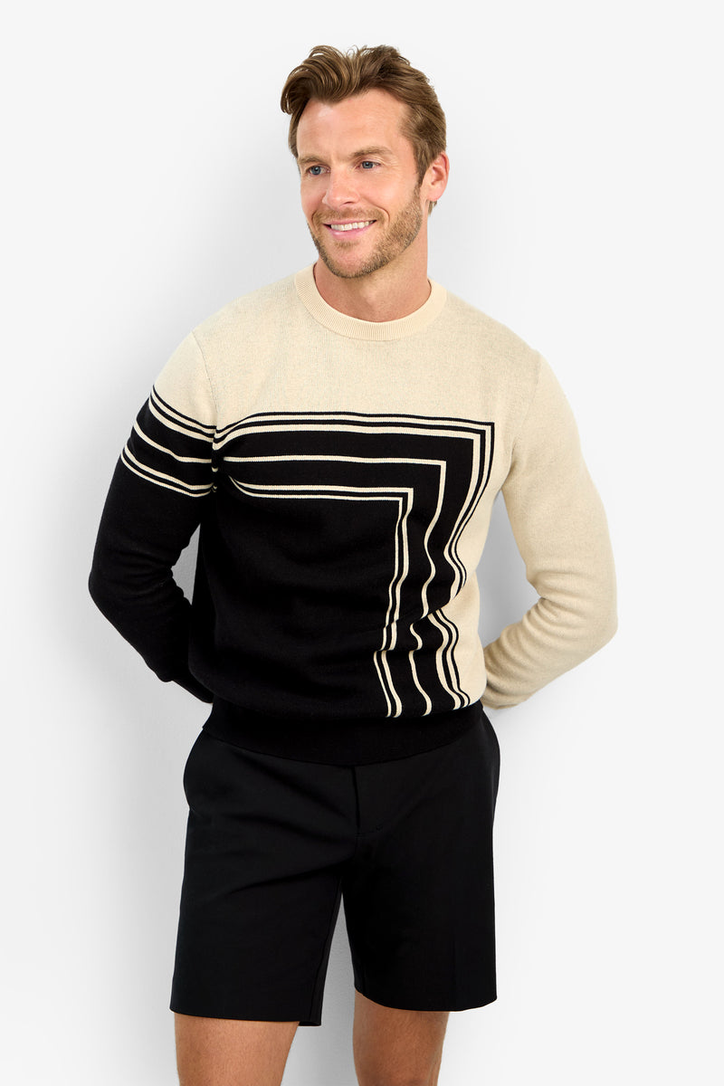 A man with light brown hair and a beard smiles, wearing the RAMOS STRIPE CREWNECK—a cream and black geometric sweater—paired with black shorts, posing against a plain light background.