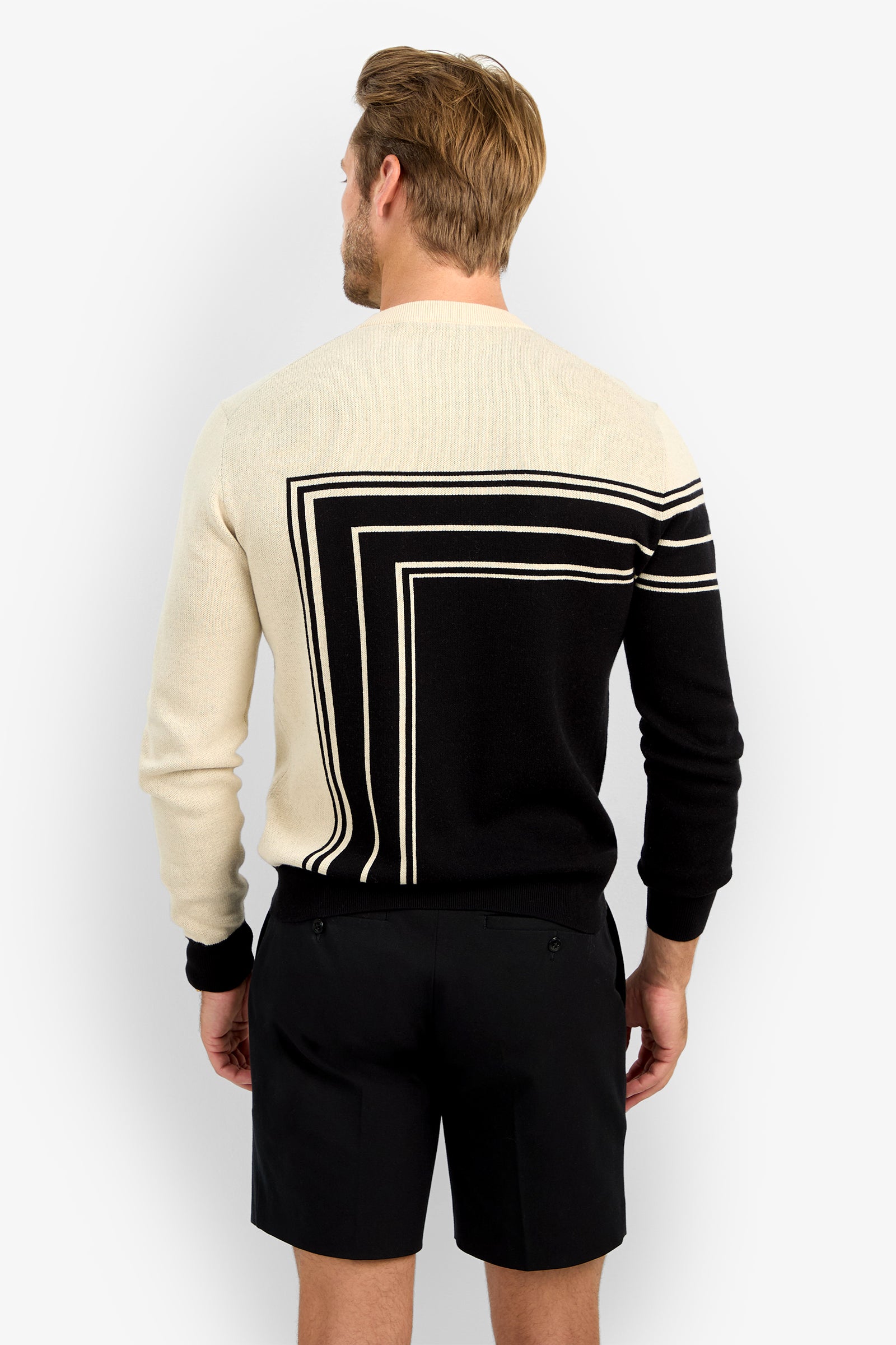 A man with light brown hair stands facing away, wearing the RAMOS STRIPE CREWNECK—a long-sleeve sweater featuring a geometric black and cream pattern on the back—paired with black shorts.