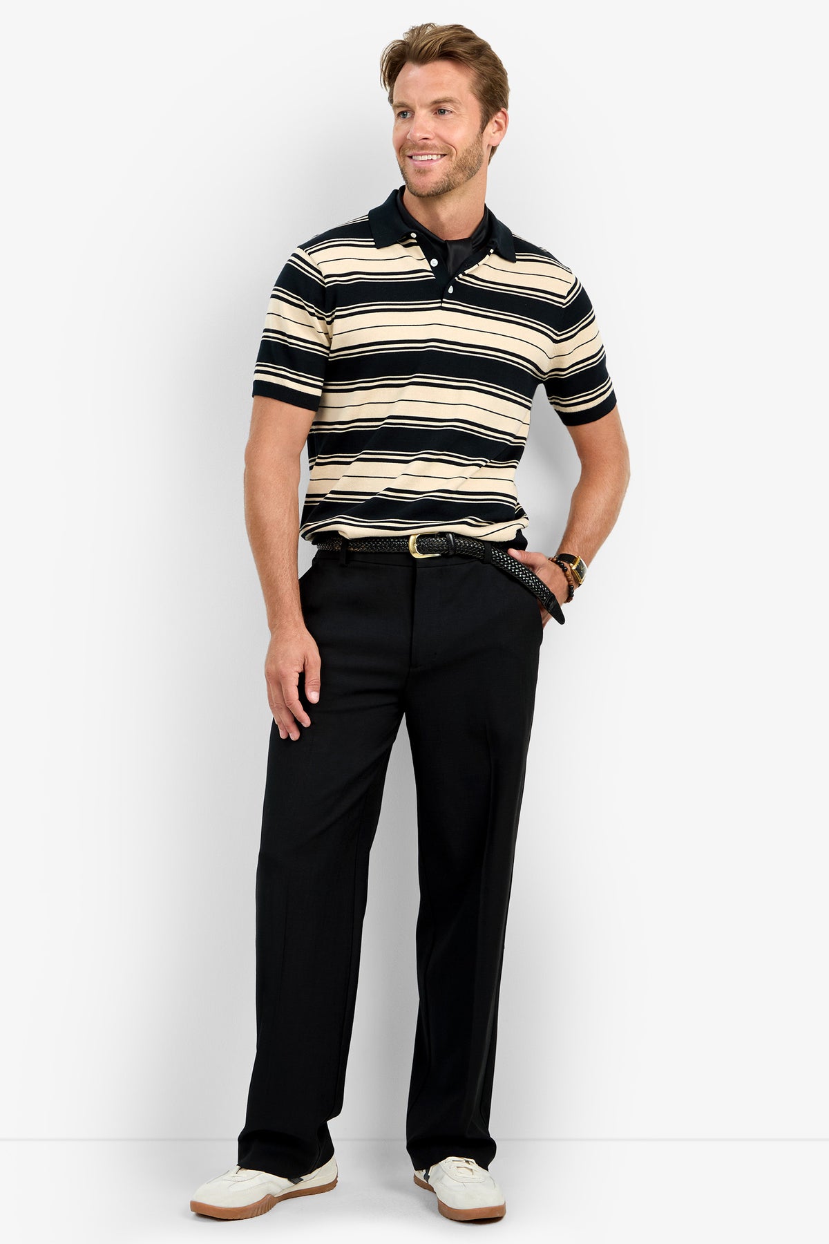 A man with short light brown hair stands smiling, his left hand in his pocket, wearing the EDWARD STRIPE POLO— a beige and black striped short-sleeve shirt—paired with black belted trousers and white sneakers.