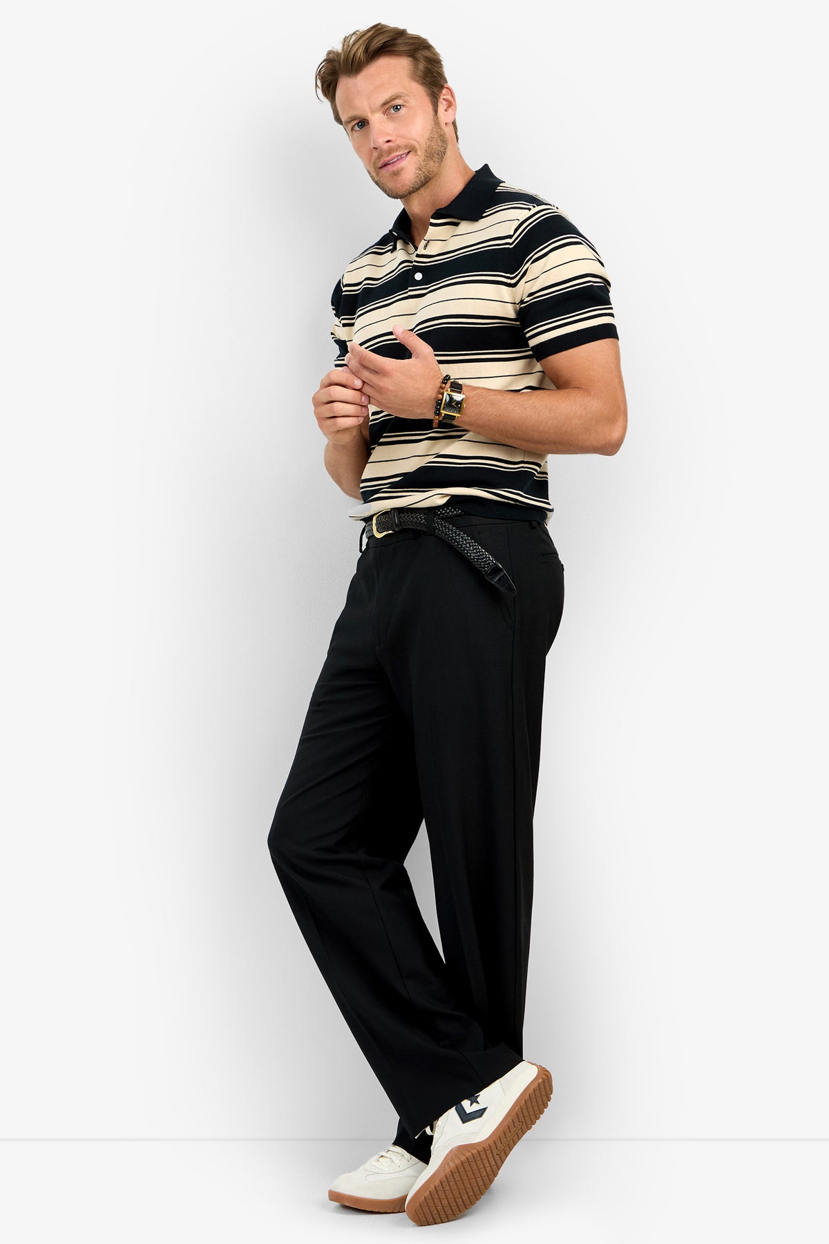 A man poses against a plain background wearing the EDWARD STRIPE POLO, black belted trousers, and white sneakers. He holds sunglasses and looks at the camera with a relaxed expression.