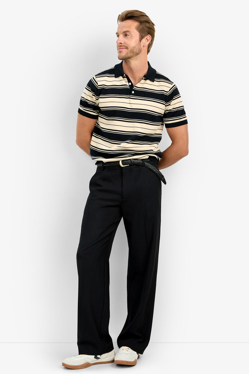 A man with short hair stands against a white background wearing the EDWARD STRIPE POLO, black belted trousers, and white sneakers. He looks to the side with his hands behind his back.
