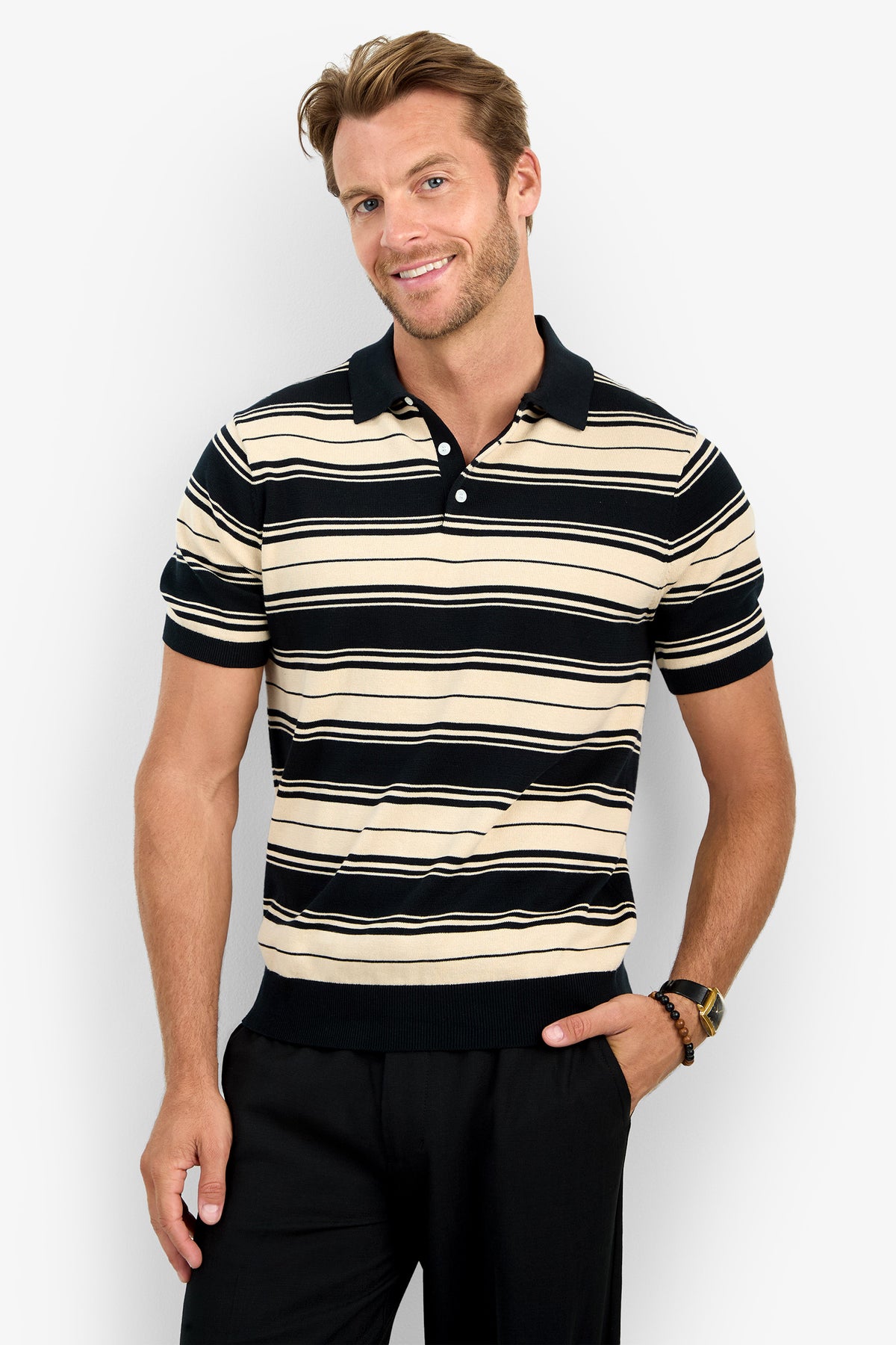 A man with short, light brown hair is smiling and posing against a light gray background, wearing the EDWARD STRIPE POLO—a short-sleeved beige and black striped polo shirt—and black pants, with one hand in his pocket.
