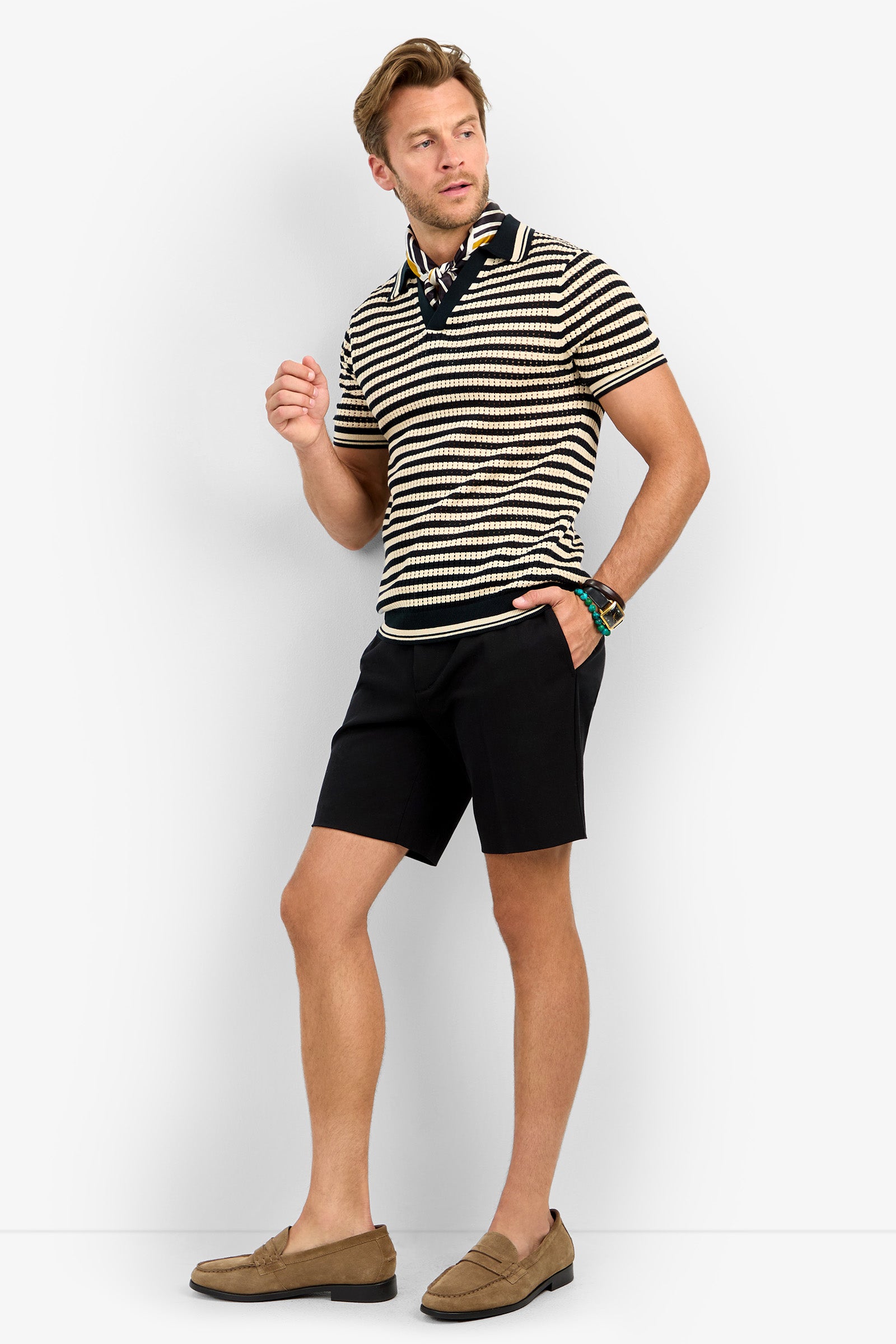 A man poses against a white background wearing the ALLEN MESH POLO, black shorts, a black neck scarf, a watch, and tan loafers. He stands with one hand raised and the other in his pocket.