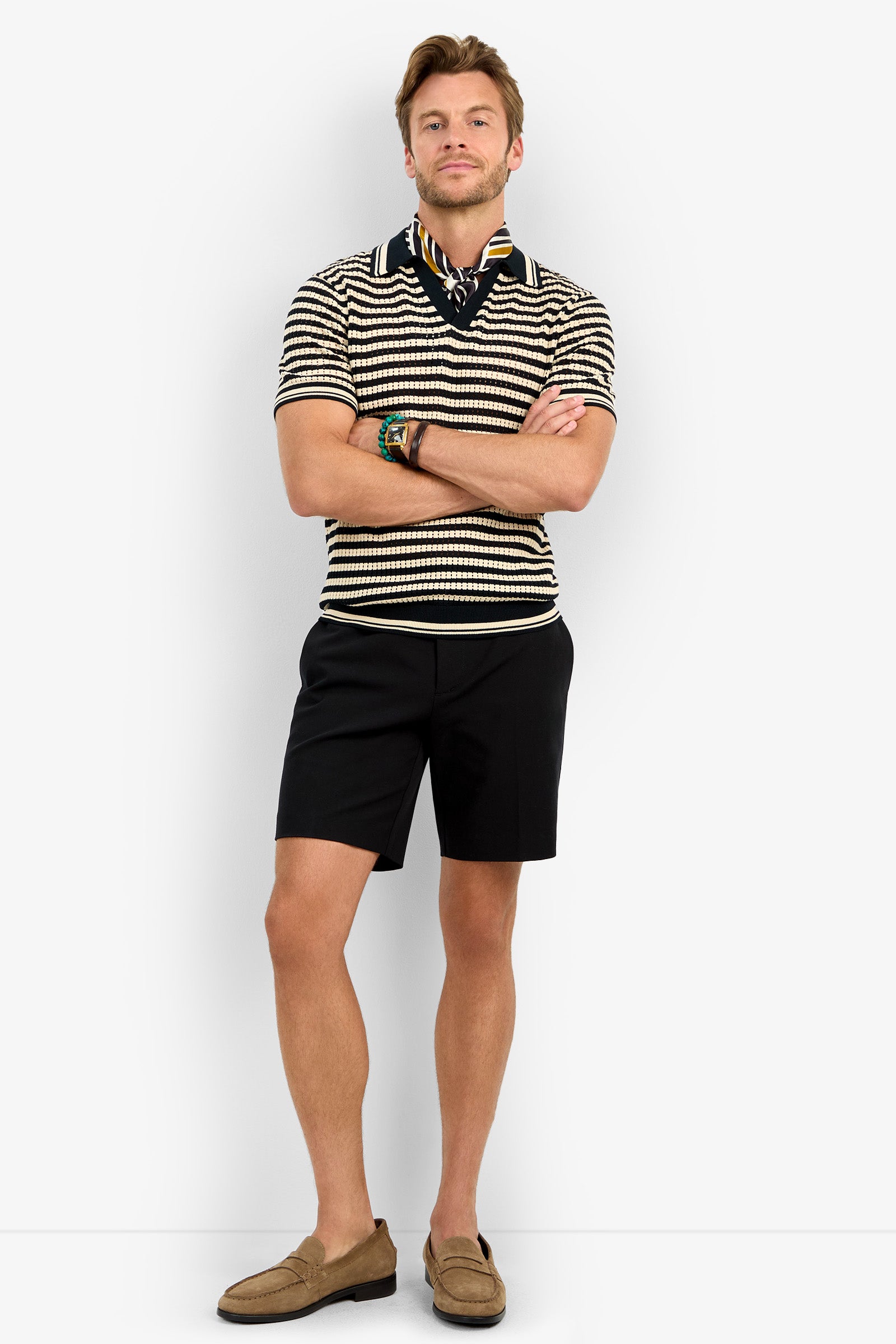 A man stands with arms crossed, wearing the ALLEN MESH POLO in black and beige stripes, paired with black shorts, tan loafers, a patterned neck scarf, and bracelets, set against a plain white background.