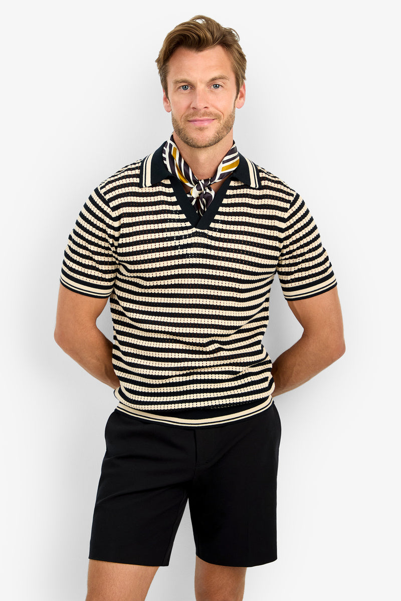 A man stands against a plain background, wearing the ALLEN MESH POLO, black shorts, and a patterned scarf tied around his neck. He smiles slightly with his hands behind his back.
