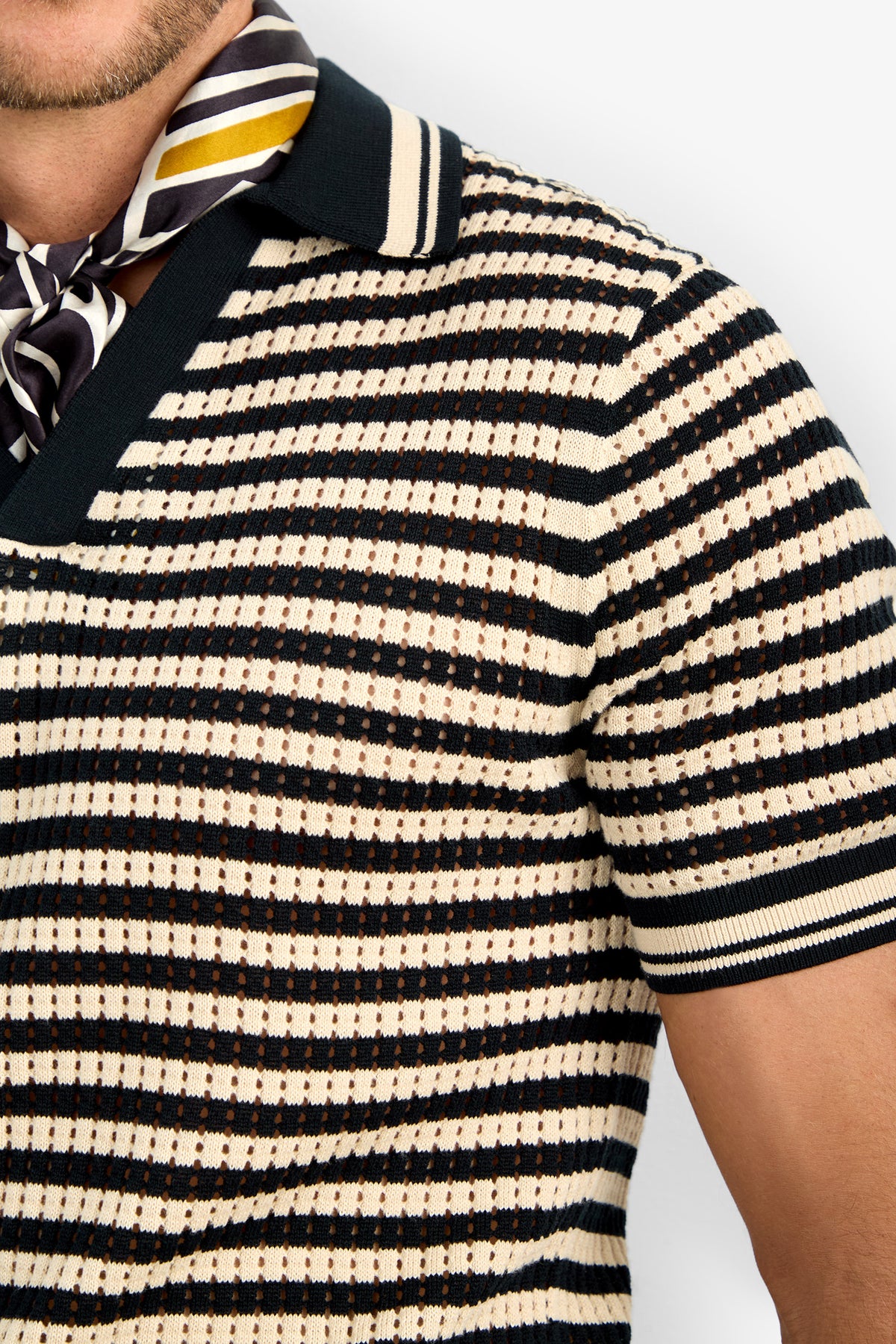 A person in the ALLEN MESH POLO, a short-sleeve striped knit shirt in cream, black, and brown with a wide collar, paired with a black, white, and yellow patterned scarf around their neck. Only their lower face, neck, and upper torso are shown.