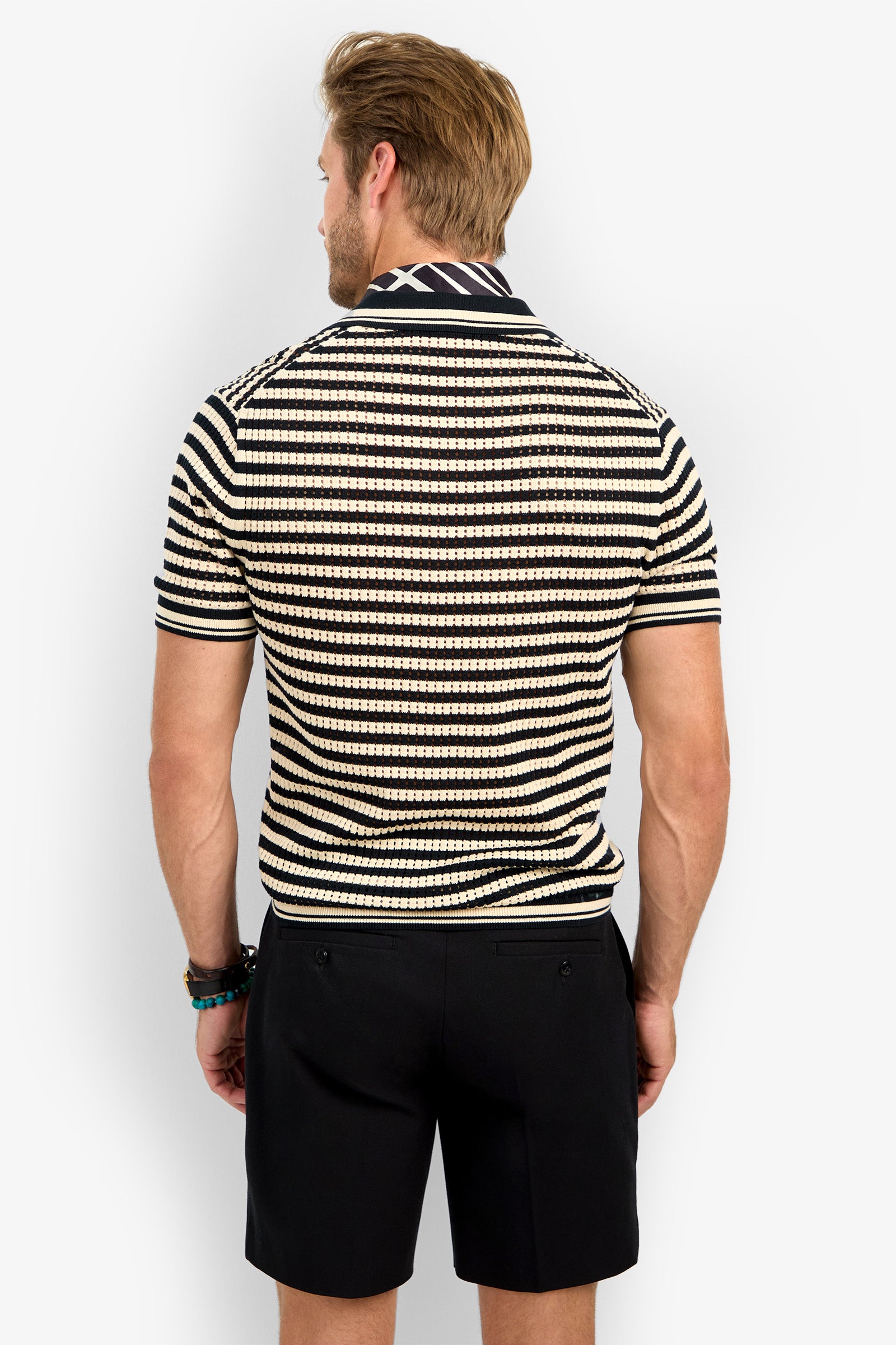 A man faces away in a beige and black striped ALLEN MESH POLO with a collar and short sleeves, paired with black shorts. His hair is styled, bracelets are on his left wrist, and the background is plain white.