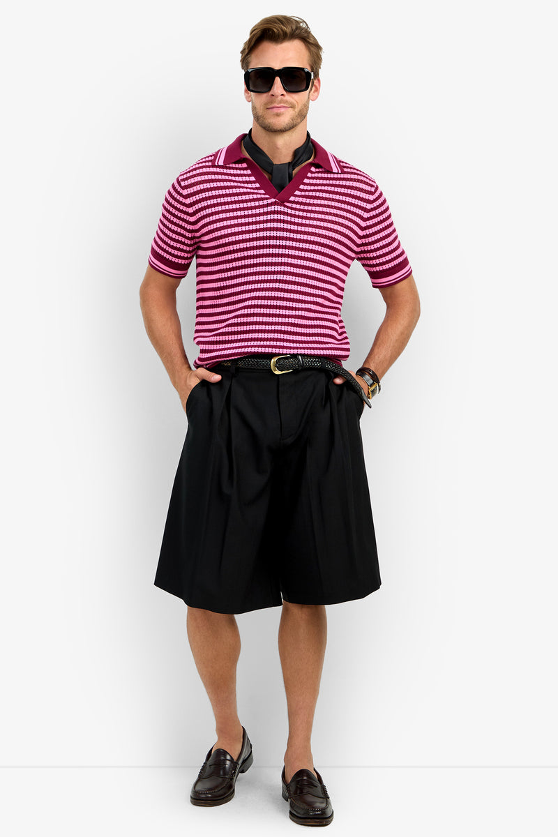 A man wearing sunglasses, an ALLEN MESH POLO in pink and white stripes, black long shorts, a black belt, brown loafers, and a watch stands with hands in his pockets against a plain white background.