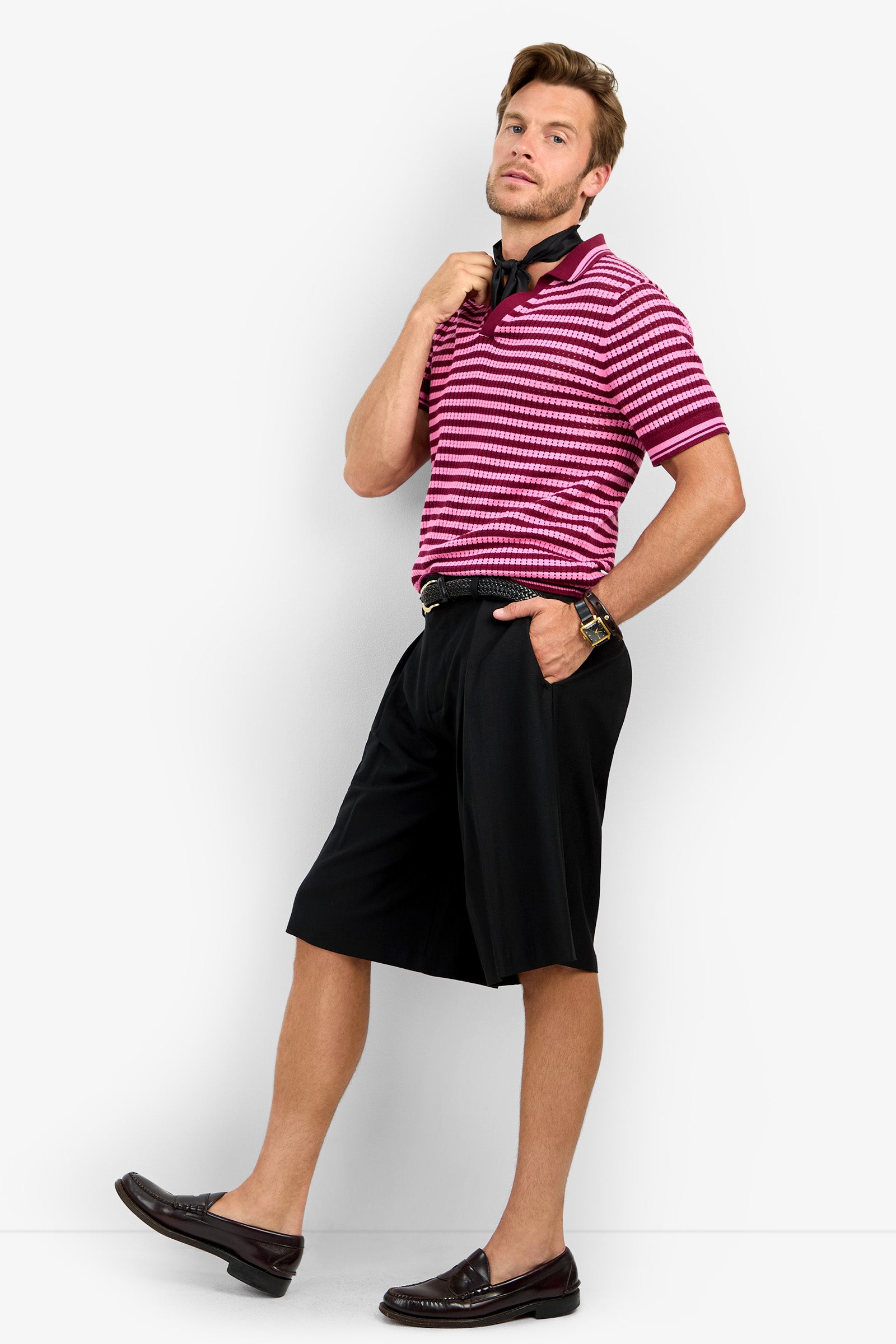 A man poses confidently in the ALLEN MESH POLO, featuring pink and white stripes, paired with black knee-length shorts and brown loafers. He has one hand in his pocket and adjusts his collar while looking at the camera.
