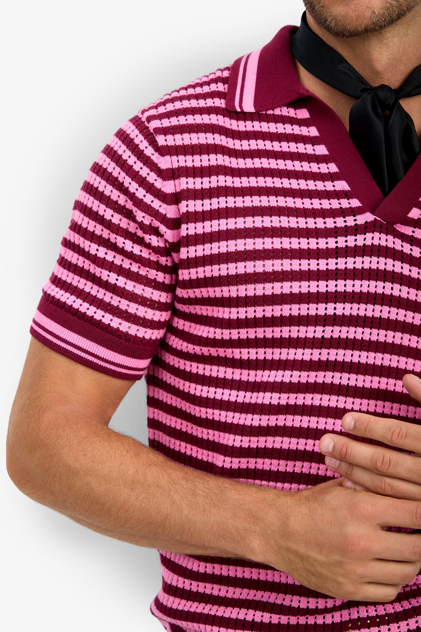 A person wears the ALLEN MESH POLO, a pink and burgundy striped collared shirt, with a black scarf tied around their neck. Their left hand is on their chest, and their face remains out of frame.