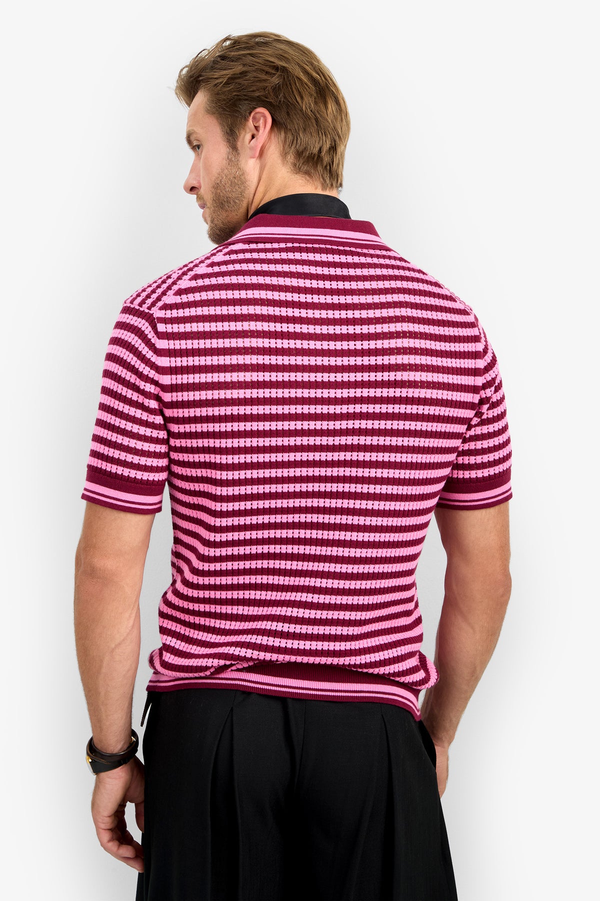 A man with light brown hair stands with his back to the camera, wearing the short-sleeved ALLEN MESH POLO in pink and maroon stripes, black pants, and a wristwatch on his left wrist.