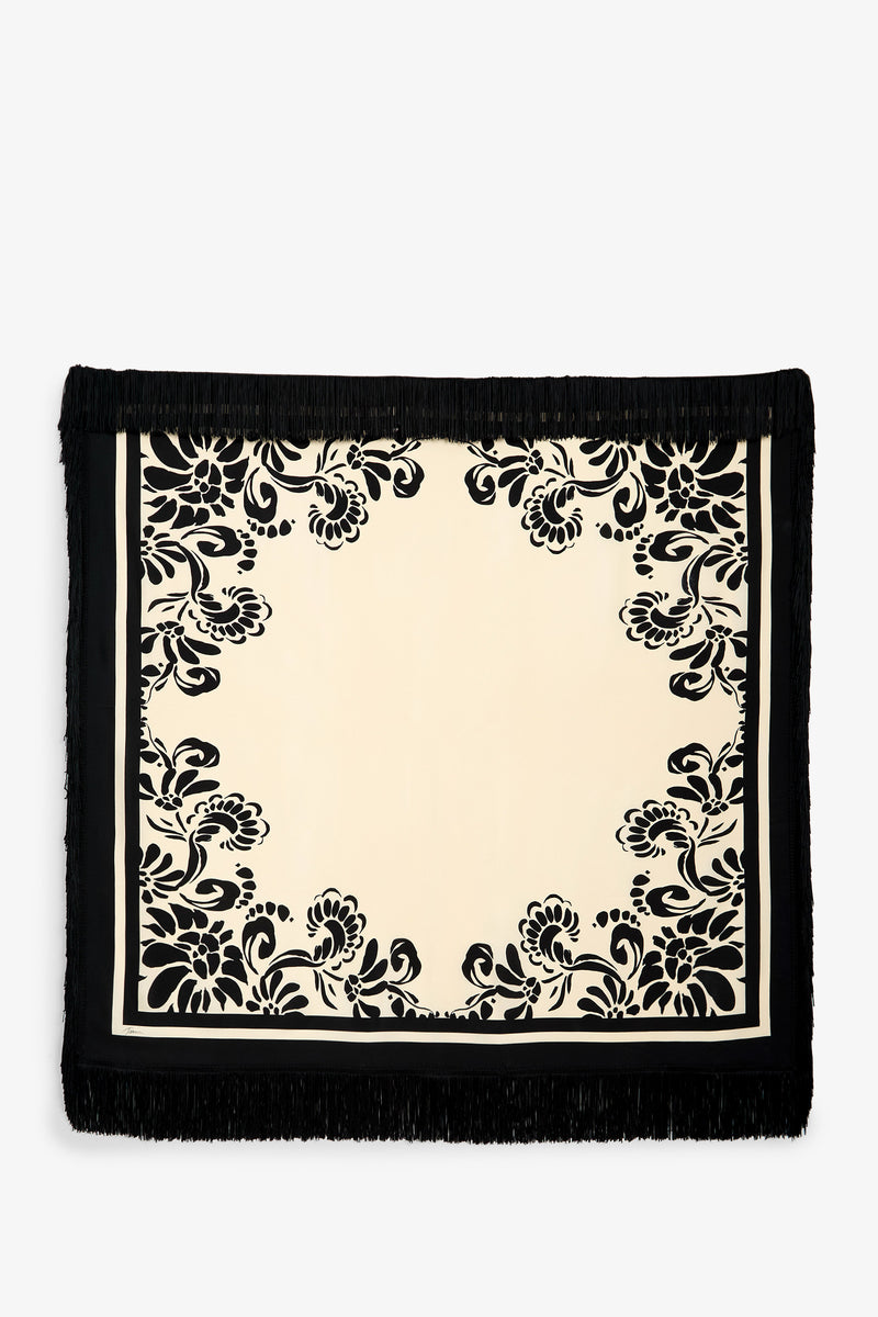 The Tivoli Fringe Scarf is a rectangular, cream-colored textile featuring black floral patterns along the border and black fringe on every side. Its center area remains blank and unadorned.