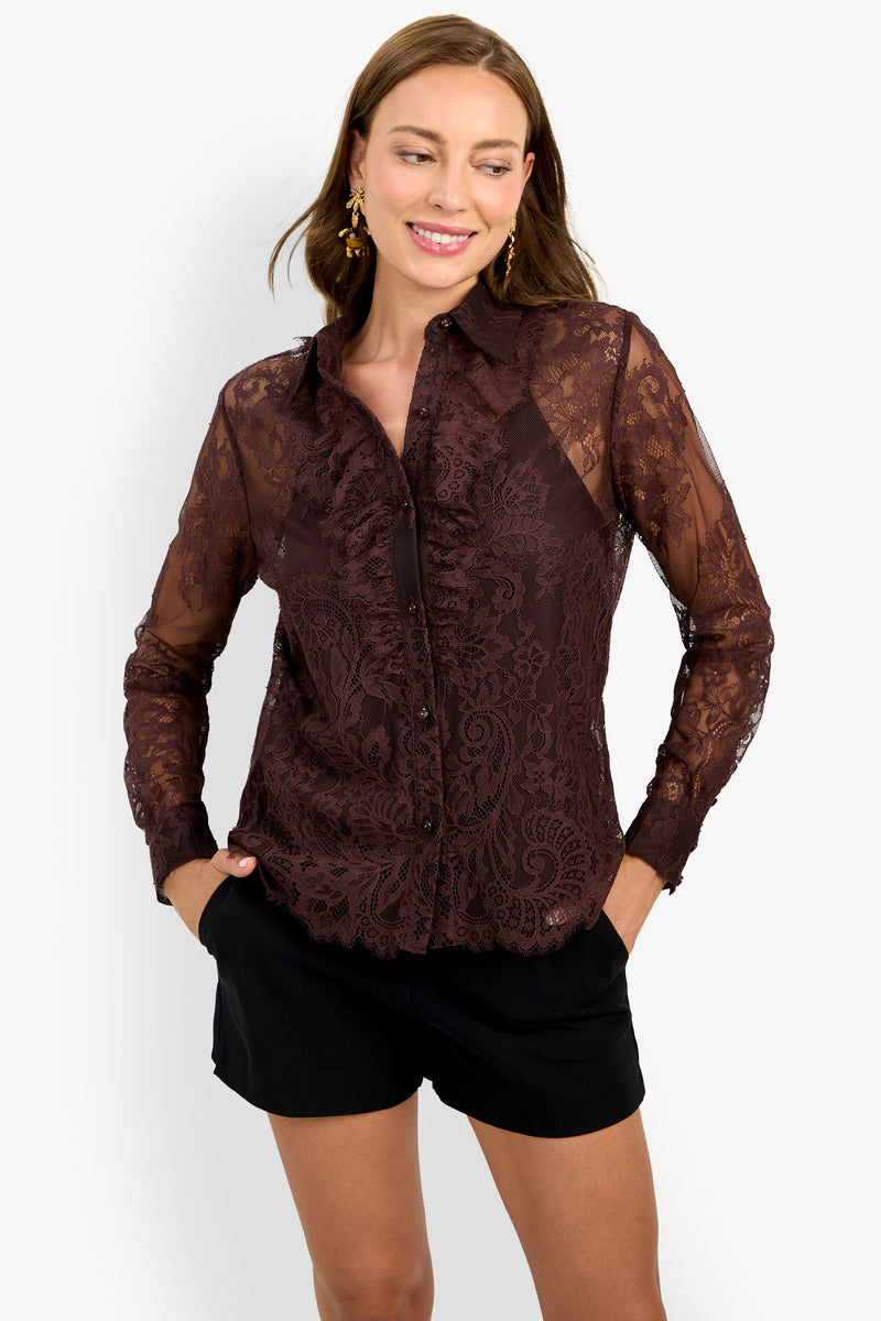 A woman models the Cassiah Top—a sheer, lace-patterned long-sleeve maroon blouse with ruffle details—paired with black shorts, smiling with her hands in her pockets against a plain light background.