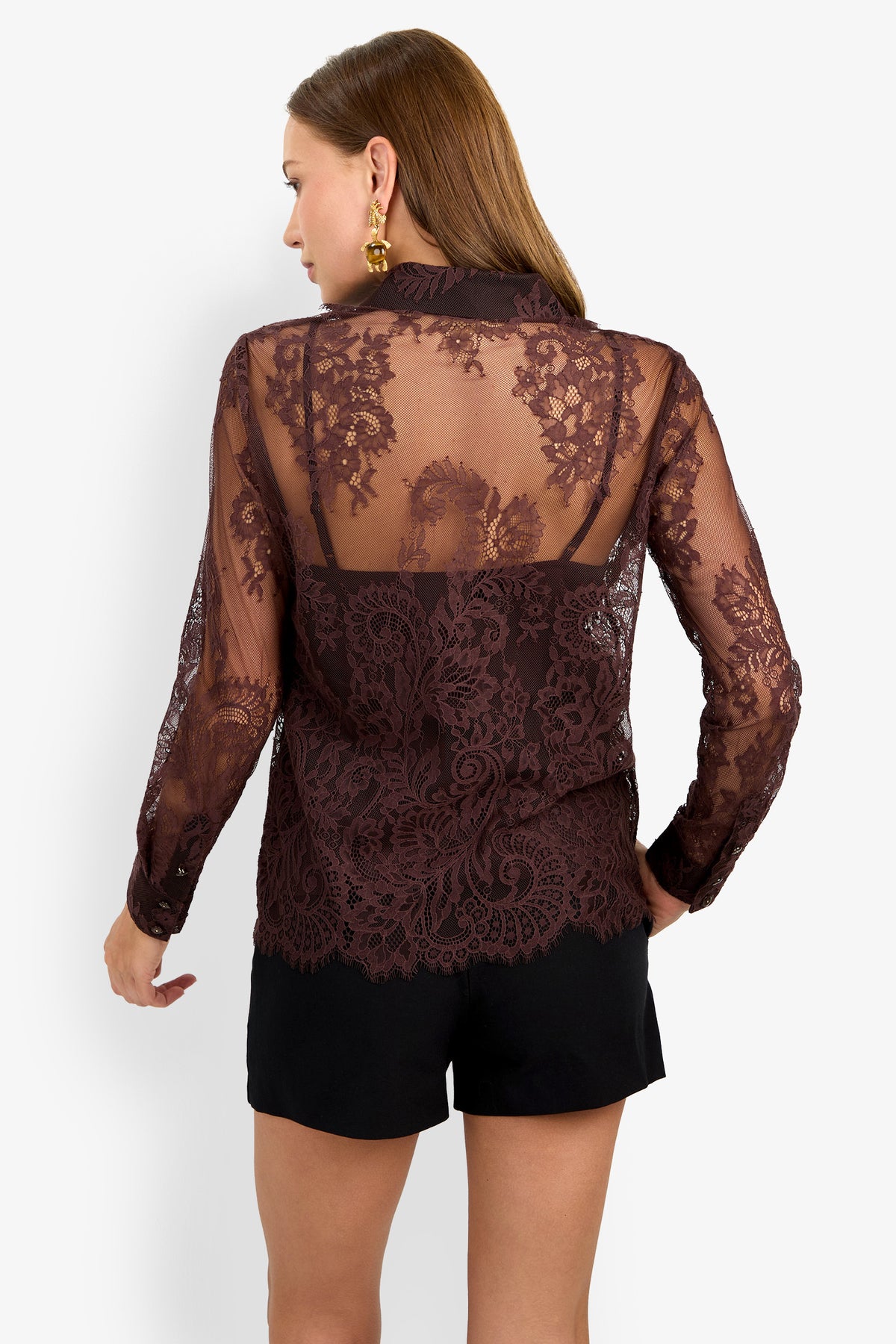A woman is seen from the back wearing the Cassiah Top, a sheer, long-sleeve blouse with intricate floral lace patterns and scalloped edges, layered over a black camisole and paired with black shorts.