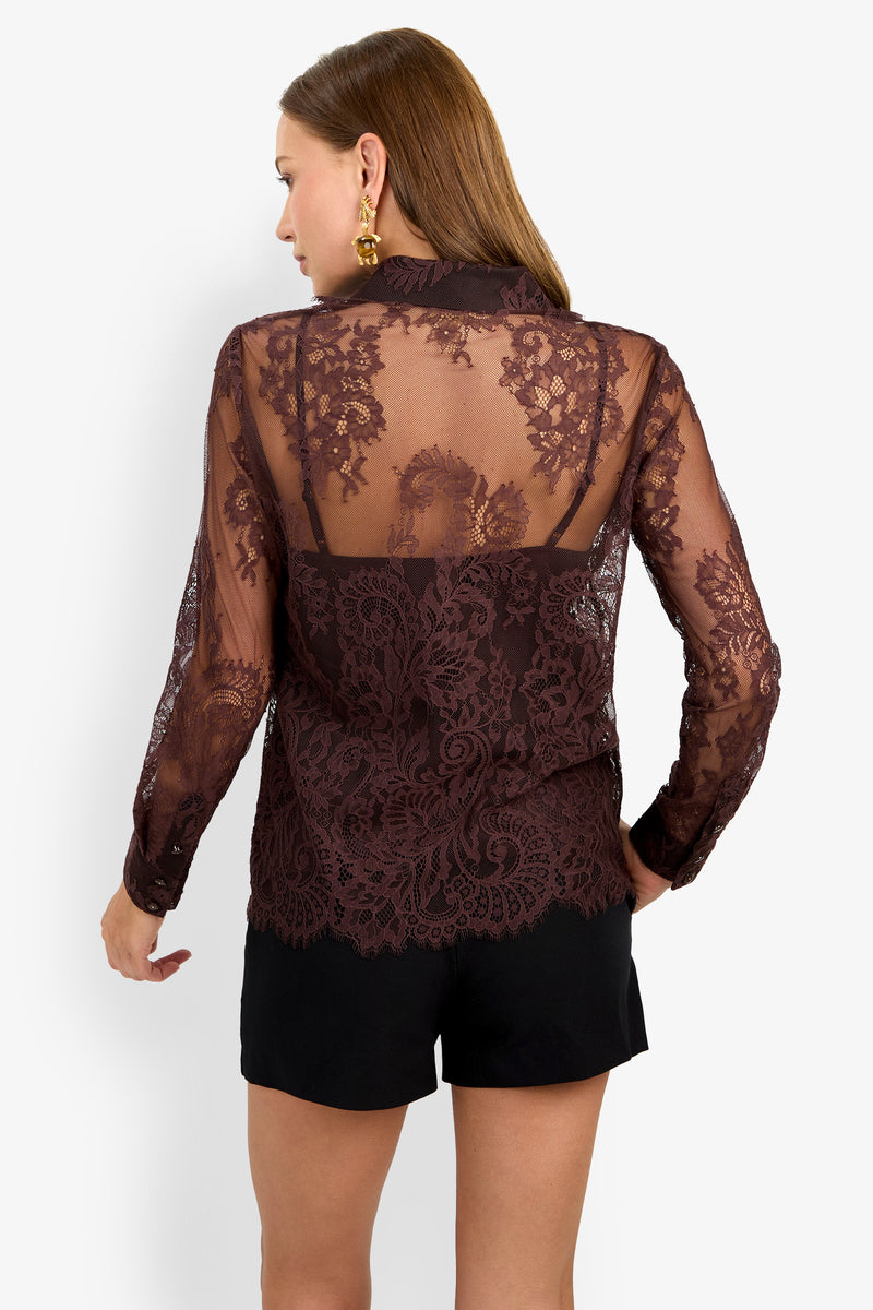 A woman is seen from the back wearing the Cassiah Top, a sheer, long-sleeve blouse with intricate floral lace patterns and scalloped edges, layered over a black camisole and paired with black shorts.