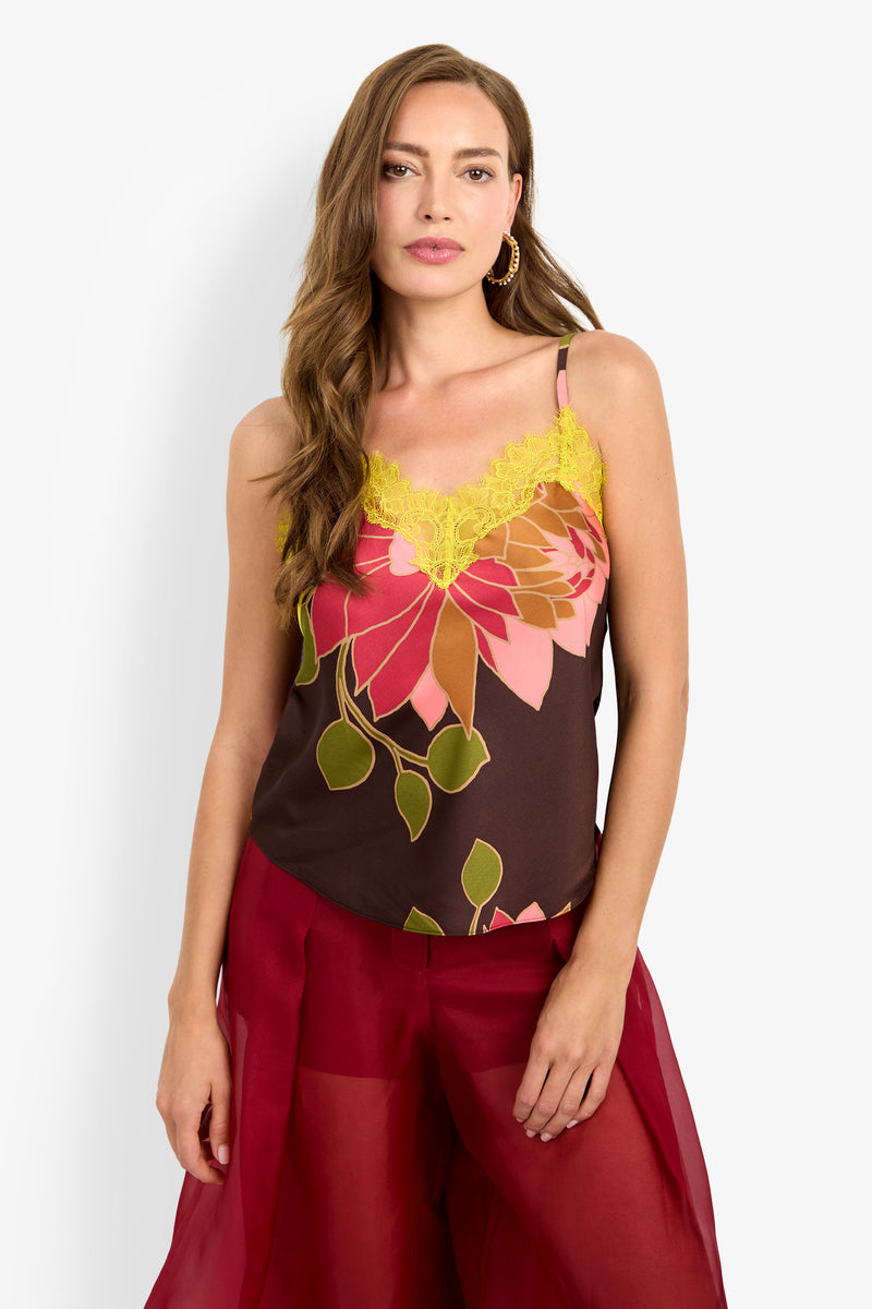 A woman with long brown hair stands against a white background, wearing the Amoira Top—a floral camisole with yellow lace trim—and dark red sheer pants.