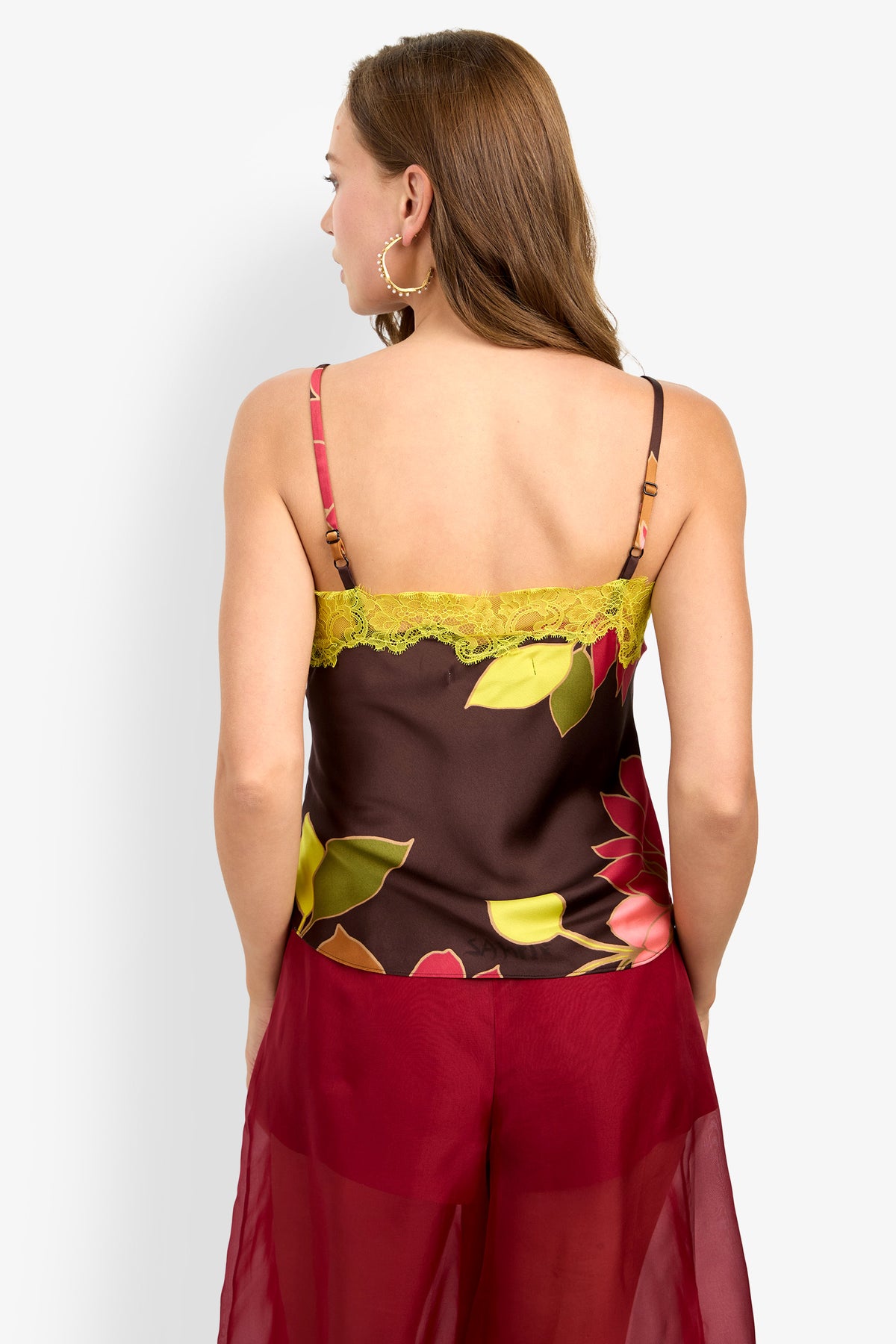 A woman with brown hair is shown from the back, wearing the Amoira Top—a floral camisole with yellow lace—and red sheer pants.