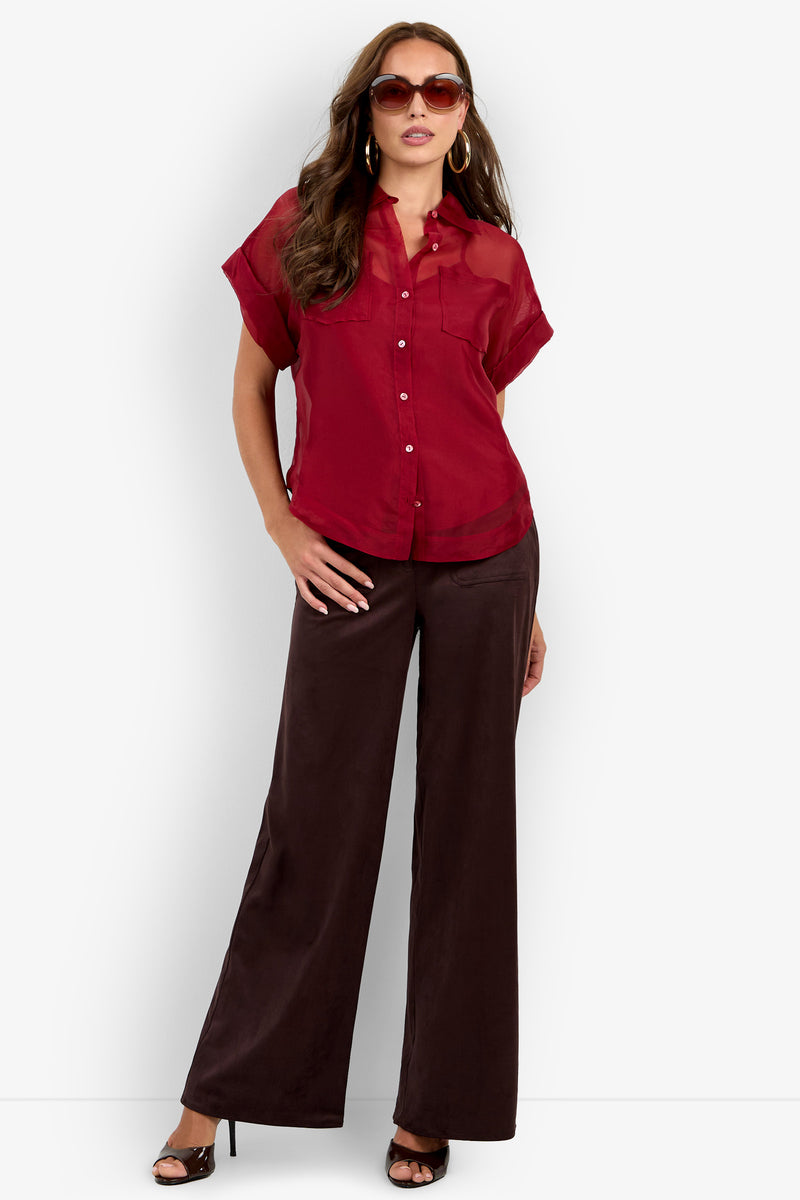 Woman wearing sunglasses, the Costanzo Top—a sheer red short-sleeve button-up blouse—and high-waisted wide-leg brown pants with black heel sandals, standing against a plain white background.