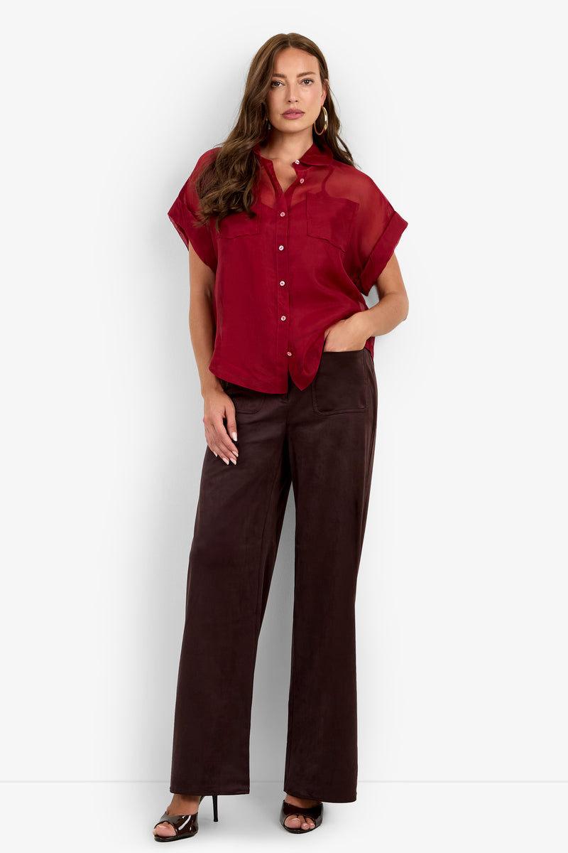 A woman wearing the Costanzo Top—a short-sleeve, button-up red blouse—paired with wide-leg dark brown pants and black heels stands against a plain white background, one hand in her pocket.