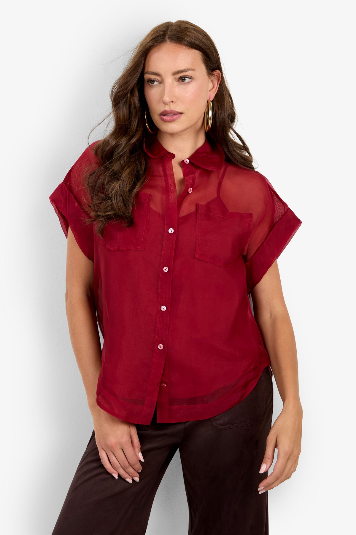 A woman wearing the Costanzo Top, a short-sleeve sheer red button-up blouse with two chest pockets, paired with dark pants. She has long, wavy brown hair and large hoop earrings.