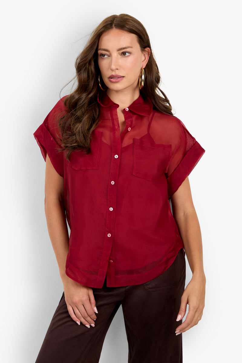 A woman wearing the Costanzo Top, a short-sleeve sheer red button-up blouse with two chest pockets, paired with dark pants. She has long, wavy brown hair and large hoop earrings.