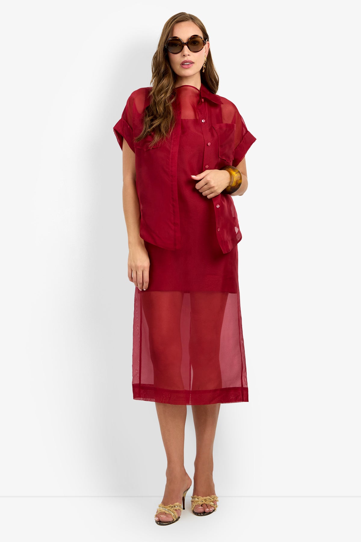 Woman wearing sunglasses, the COSTANZO TOP—a sheer red short-sleeve shirt—layered over a matching red sheer midi dress, yellow heeled sandals, and a chunky brown bracelet, standing against a white background.