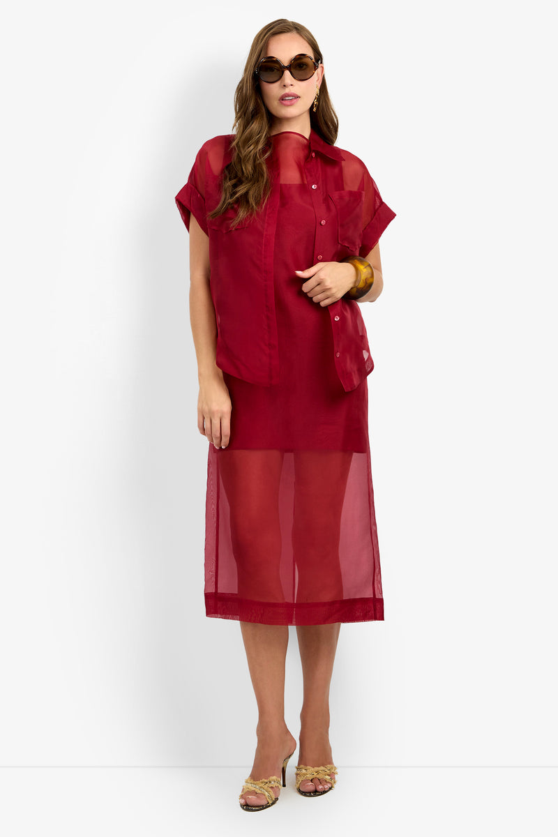 Woman wearing sunglasses, the COSTANZO TOP—a sheer red short-sleeve shirt—layered over a matching red sheer midi dress, yellow heeled sandals, and a chunky brown bracelet, standing against a white background.