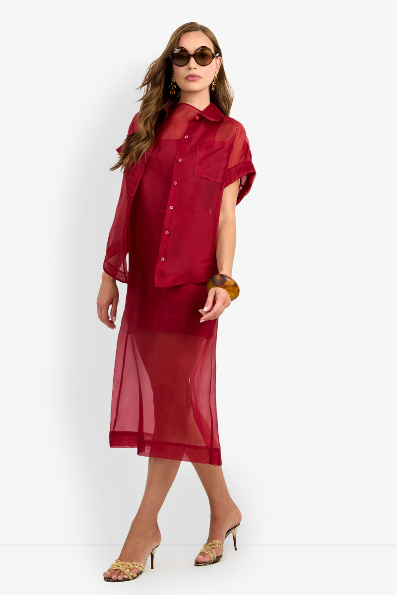 A woman poses against a plain white background wearing the sheer red COSTANZO TOP over a matching dress, styled with sunglasses, a brown bangle, and heeled sandals.